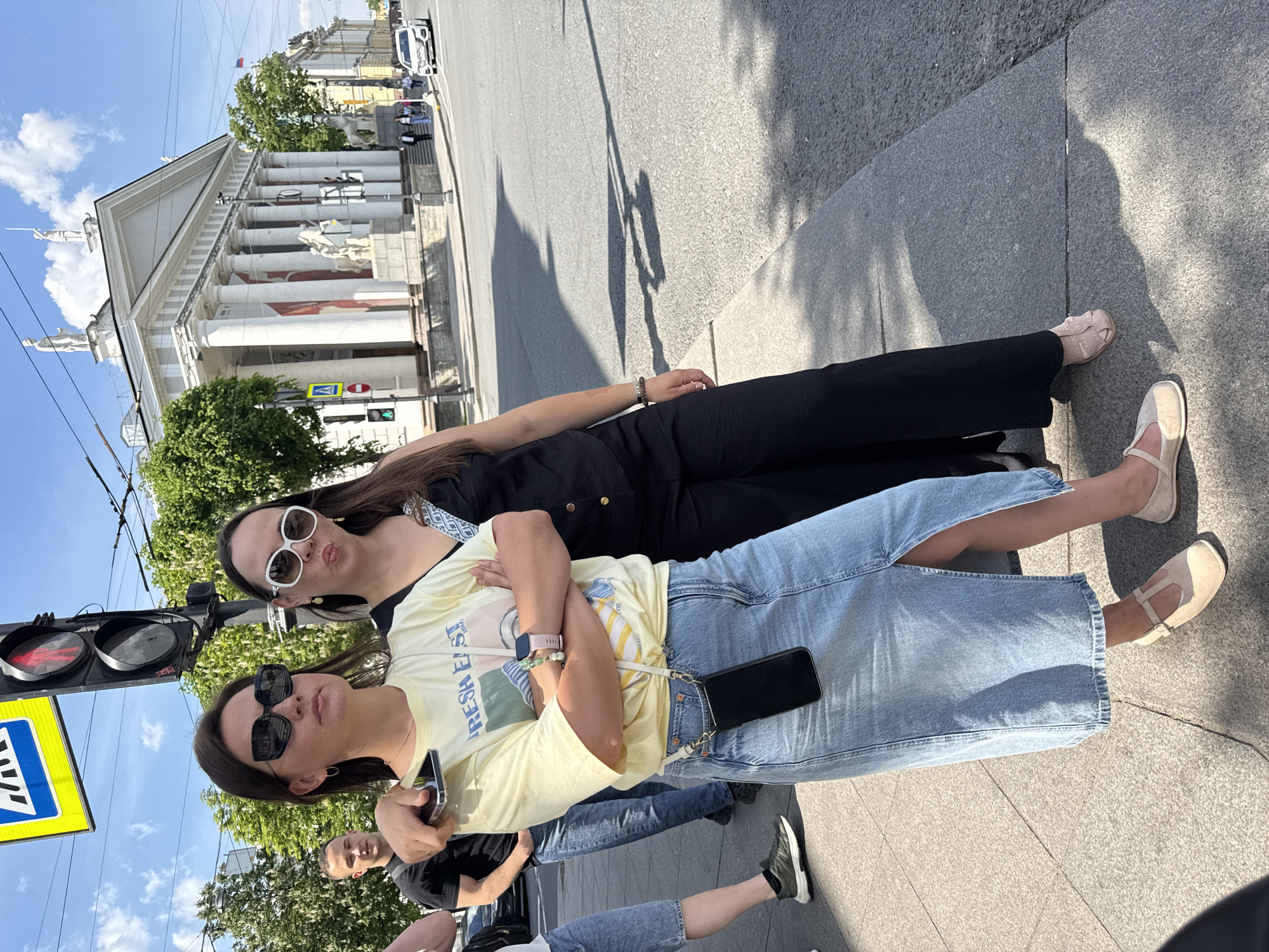 The image, rotated 90 degrees clockwise, captures two young women posing on a sunny city street in what appears to be Saint Petersburg, Russia. Behind them stands a grand, white classical building featuring numerous tall columns, a triangular pediment, and statues along its roofline and facade.
The woman on the left wears a light yellow t-shirt with the text "FRESH EAST" printed on it, a light blue denim midi skirt with a side slit, and light-colored flat shoes. She has dark hair, wears black sunglasses, and holds a black smartphone in her hand, with another phone attached to her skirt by a strap. Her arms are crossed.
The woman on the right wears a black jumpsuit or dress, white-framed sunglasses, and light-colored flat shoes. She has dark hair and is making a playful "duck face." Partially visible behind the women are the face and leg of another person.
The setting includes a paved sidewalk in the foreground, an asphalt street beyond, and green trees. To the left, a black traffic light pole with red and green lights is prominent, alongside a blue square pedestrian crossing sign. The sky is blue with scattered white clouds, and long shadows are cast on the ground, indicating bright sunshine during daytime.