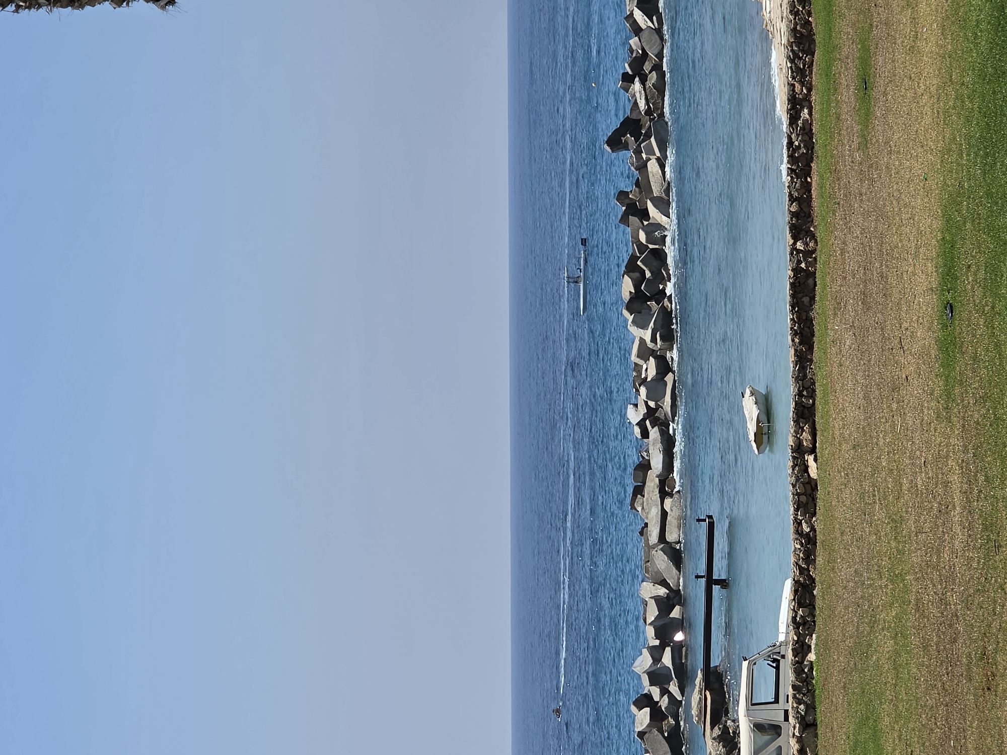 The image shows a coastal scene in Hlorakas Community, Cyprus, on a clear, sunny day. The foreground features a patch of dry, golden-brown grass interspersed with greener patches, bordering a low, stone wall. Behind the wall, a white jeep is parked on a paved area next to the water. A small, white boat is moored in the calm, clear turquoise water. Further out, a breakwater made of large, angular concrete blocks separates the calm bay from the open sea. The sea is a deep blue, with gentle waves lapping at the breakwater. On the horizon, the vast expanse of the sea meets a clear, light blue sky. In the distance on the water, a person on a jet ski is visible on the left, leaving a white wake, and a small boat is cruising on the right.