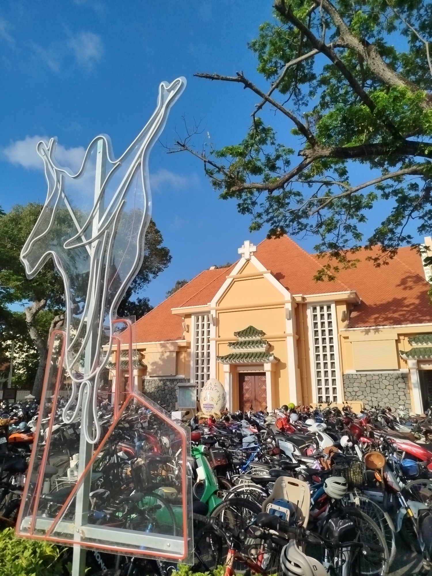 A photo captures the outside of a church in Ho Chi Minh City, Vietnam on a clear, sunny day. The church is characterized by its orange tiled roof, a cross at the apex, and light yellow walls. A multitude of bicycles and motorbikes are parked densely in front of the building, occupying much of the foreground. A modern, transparent art installation stands prominently to the left of the frame, featuring elongated figures with raised arms. The sky above is a bright blue with scattered clouds, and the upper right corner is framed by the branches of a tree with green leaves.