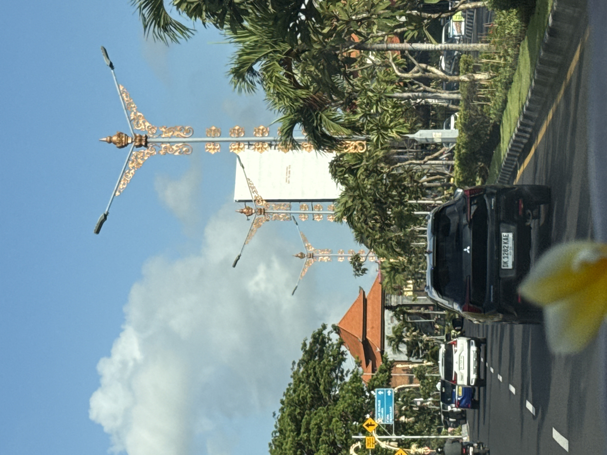 This image captures a street scene in Kuta, Indonesia, likely during the day under a clear sky with some clouds. The foreground shows the rear of a black SUV driving on a paved road. The SUV has a license plate visible, reading "DK 1282 KAE". In front of the SUV, other vehicles, including a white SUV and a blue car, are also on the road. A person on a motorcycle is seen in the left lane.
The roadside is lined with palm trees and other lush green vegetation. Elaborate streetlights with ornate golden decorations adorn the sides of the road, suggesting a well-maintained or possibly a festive area. In the background, a building with a traditional orange-tiled roof is visible, along with a large white billboard or structure. Traffic signs, including directional arrows and warnings, are also present. The overall environment suggests a tropical urban setting with ongoing vehicular traffic.