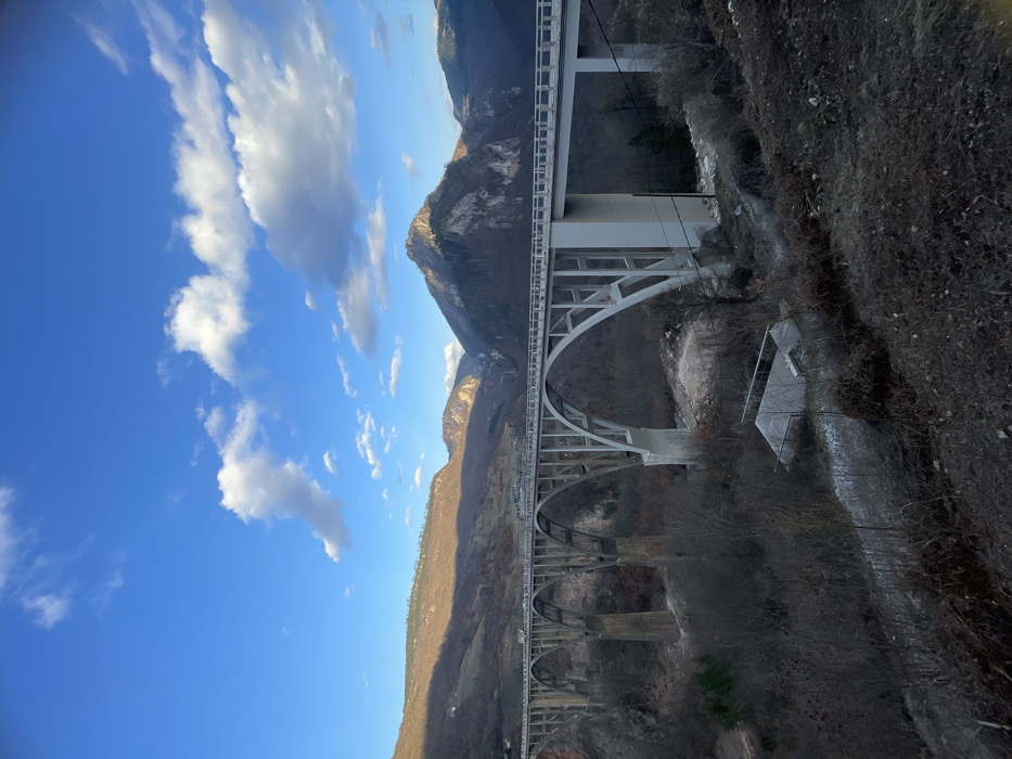 The image displays the Đurđevića Tara Bridge in Đurđevića Tara, Montenegro, a multi-arch concrete bridge spanning a deep, forested river canyon. The light grey bridge features five prominent arches supporting a flat roadway where several vehicles are visible as small light-colored forms. The surrounding landscape consists of rugged mountains under a clear blue sky with scattered white cumulus clouds. Hills on the left exhibit sparse, brown deciduous vegetation under partial sunlight, while mountains to the right and in the background are darker, showing exposed rock faces and patches of evergreen forest. Beneath the bridge, the canyon floor is covered with dormant, dark-branched trees and shrubs. A small, rectangular building with a light-colored roof is situated on the slope below the right section of the bridge, near dark utility lines descending the hillside and a visible dirt path. No individuals are discernible in the scene.