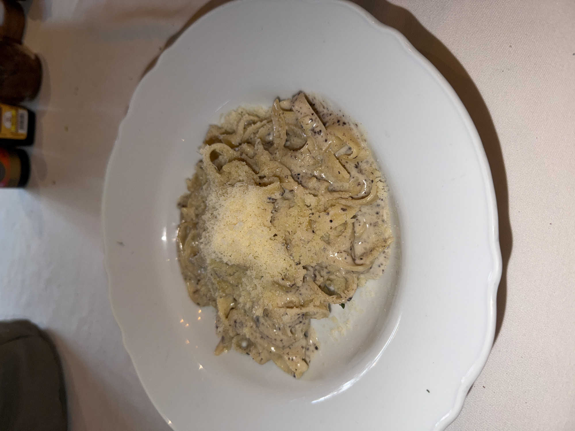 A plate of pasta is centrally located on a white, ruffled-edged plate. The pasta appears to be fettuccine or tagliatelle, coated in a light-colored, creamy sauce with dark specks. A mound of grated cheese covers a portion of the pasta. To the left of the plate are blurry objects, including what appears to be a bottle. The plate rests on a neutral-colored tablecloth. The location is inferred to be Domingo Arenas, Mexico.