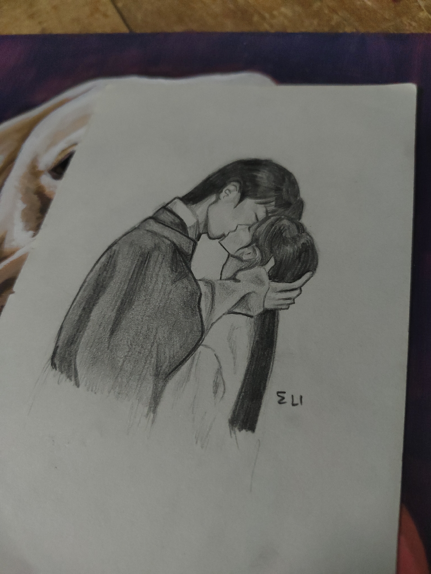 A pencil drawing on a white sheet of paper depicts two individuals, likely a man and a woman, engaged in an embrace and a kiss. The drawing is rendered in shades of grey, indicating pencil as the medium. The individual on the left, presumably male, is taller and wears a dark, long-sleeved garment with a prominent collar. His head is tilted down towards the other individual, and his hair appears dark and styled. The individual on the right, presumably female, has long, dark hair extending down her back. Her head is tilted upwards, and her face is partially obscured as she meets the kiss. Her right hand is visible, gently cupping the back of the male figure's head. Both figures appear to be wearing traditional or stylized robes, suggesting a possible historical or fantasy context. The initials "ELI" are written vertically in capital letters at the bottom right corner of the drawing.
The paper itself is resting on a patterned surface, possibly a desk mat or mousepad, visible beneath the paper's edges. This surface features shades of dark blue or purple, with hints of a lighter, possibly tan or off-white, element in the top left corner that vaguely suggests part of an animal's face or fur. To the top right, a wooden or textured surface is visible.
There is no discernible visual information within the image that allows for the identification of a specific location in Davao City, Philippines.