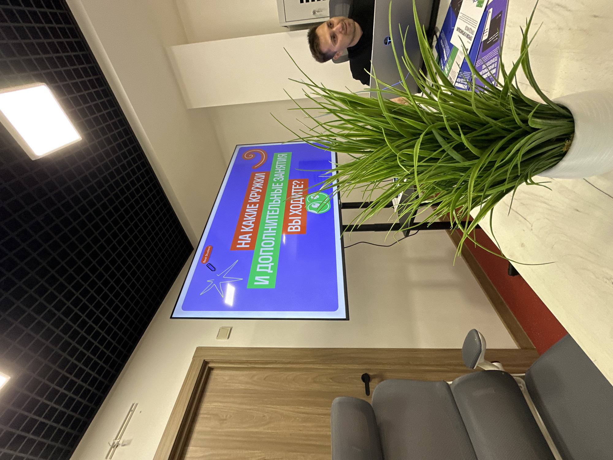 This media file depicts an indoor scene, likely an office or classroom, in Sochi, Russia. The main subject is a large screen displaying text and graphics in Russian. The text on the screen reads "НА КАКИЕ КРУЖКИ И ДОПОЛНИТЕЛЬНЫЕ ЗАНЯТИЯ ВЫ ХОДИТЕ?" which translates to "What clubs and extracurricular activities do you attend?". There are also graphical elements such as a spiral, a starburst, and icons of a pencil and a smiley face with a green background.
In the foreground, a large potted artificial plant with vibrant green leaves partially obscures the screen. To the left of the screen, a dark grid-patterned ceiling with illuminated panels is visible, suggesting artificial lighting. The background shows a portion of a doorway with a wooden frame, and on the right side, a man in a black shirt is seated at a desk, looking towards the screen. He is partially visible behind the plant. Behind him, there is a laptop and some documents with visible text, including "ТЕХНОЛО" and "ПРАЗДНИКОВ", which are Russian words. The overall lighting suggests it is daytime, but the indoor setting makes it difficult to determine the exact time of day or weather conditions outside. The scene appears to be a moment during a presentation or a casual discussion, with the man possibly being an instructor or participant.