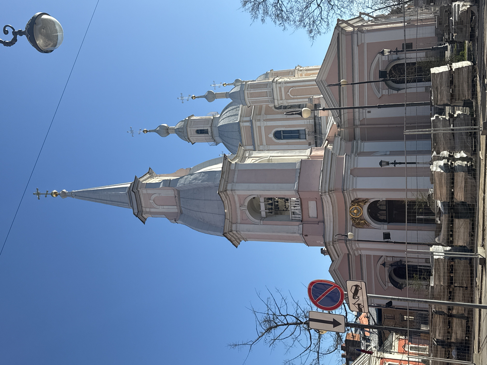 The image features the Church of Saint Catherine in Saint Petersburg, Russia. The building is primarily pink and white, with grey domes and spires topped with crosses. Construction or renovation work is ongoing, indicated by the presence of covered materials and wire fencing along the base of the building. Street signs are visible near the base of the church, including a "no stopping" sign and a directional arrow. A street lamp with a round, glass fixture is visible on the left side of the frame. The sky is clear and blue, with bare tree branches scattered throughout the upper portion of the image.
