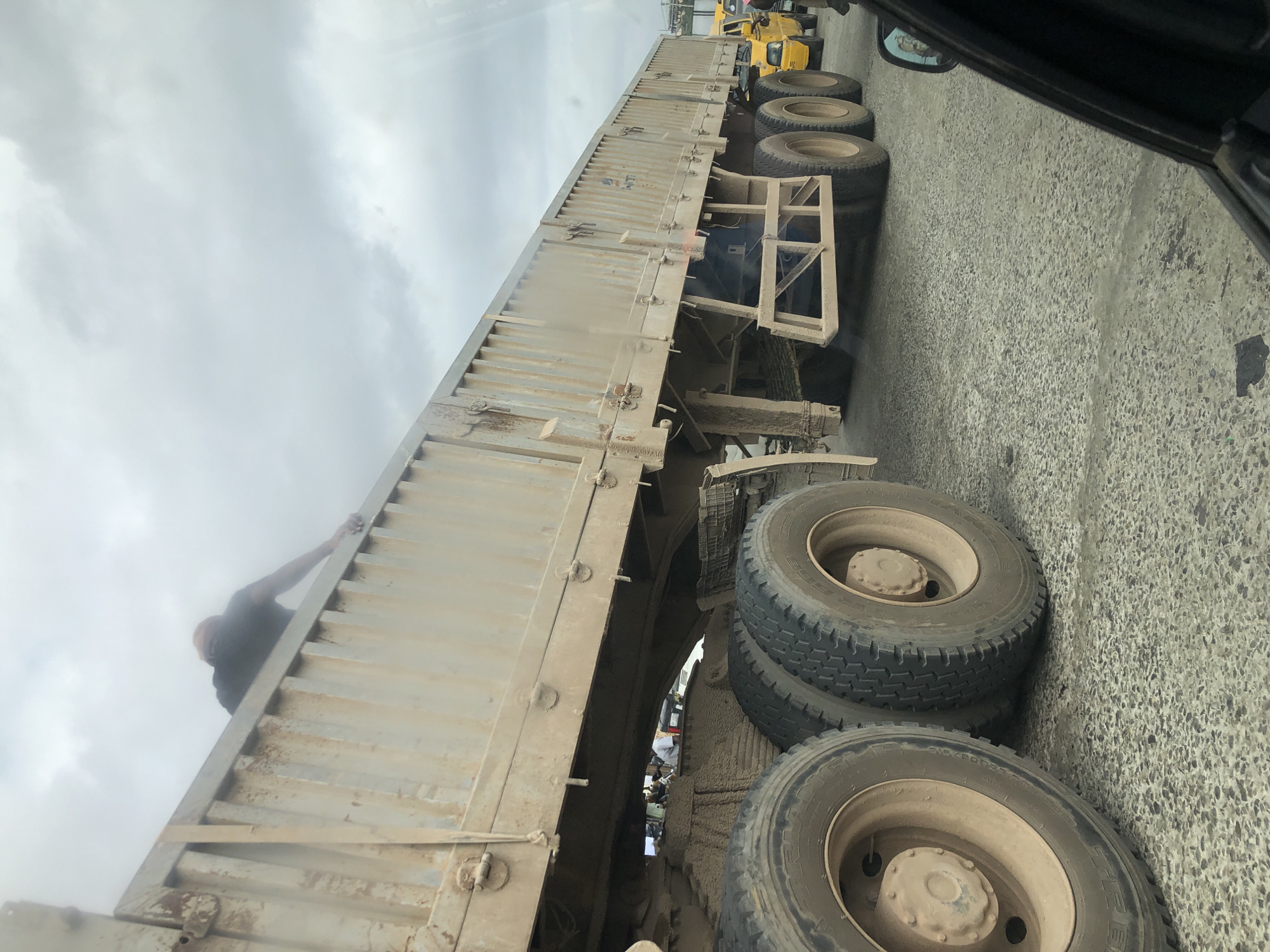 An outdoor, eye-level shot taken from inside a vehicle looking towards a large, dirty, beige-colored semi-trailer truck in traffic. The truck is very long and appears to be carrying some type of cargo, though it is not clearly visible. A person, wearing a dark t-shirt and what appears to be a light-colored hat or hood, is leaning over the top edge of the truck's trailer, with their right arm extended and hand touching the edge of the trailer. Their body is angled towards the inside of the truck bed. The truck is on a paved road, and other vehicles, including a yellow taxi and another truck, are visible in the background. The sky is overcast with clouds. The scene is likely in Otta, Nigeria, a busy industrial city.