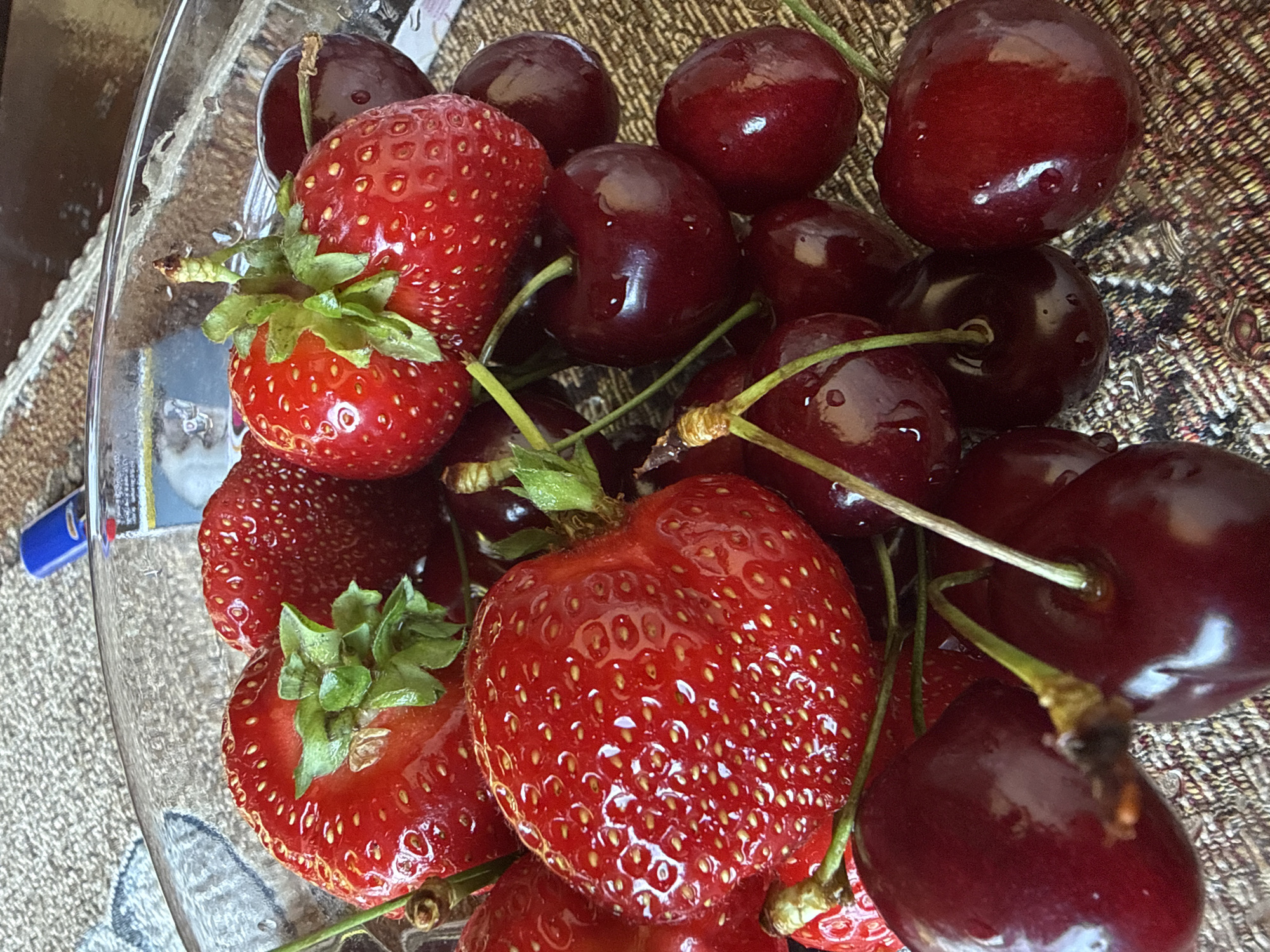 A close-up, top-down shot captures a clear glass bowl filled with fresh strawberries and dark red cherries, resting on a textured, woven placemat. The fruits are glistening, suggesting they are recently washed. Some strawberries have green leafy tops. The lighting is bright, highlighting the vibrant red of the fruits and the reflective surfaces of the bowl and the water droplets on the cherries. A blue lighter is visible in the upper background. The scene is likely in Rudartsi, Bulgaria, given the context of the location.