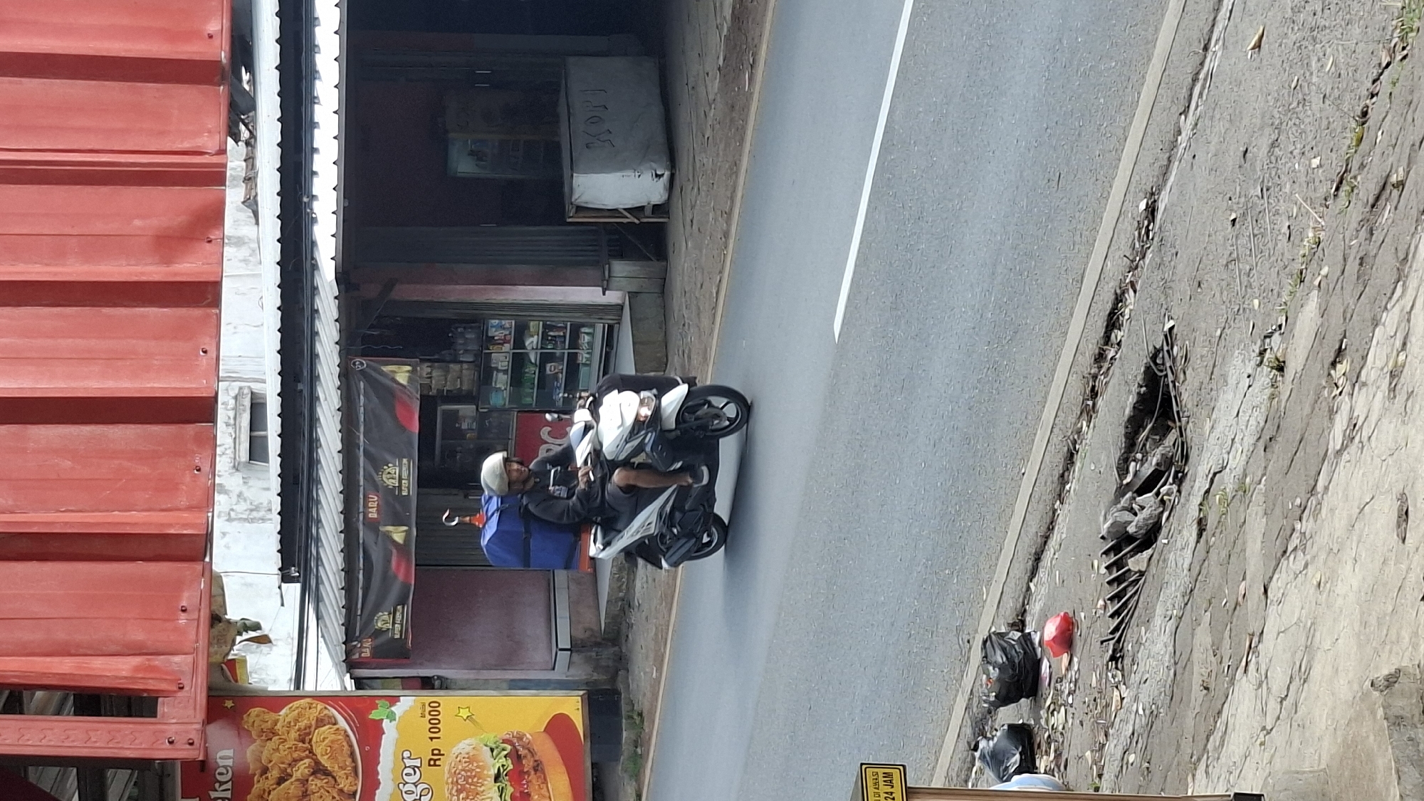 A full-shot, eye-level, outdoor, daytime photograph shows a male food delivery driver on a white scooter traveling on a paved road in Cilaku, Indonesia. The man, wearing a dark shirt, dark shorts, and a light-colored helmet, has a large blue delivery box secured to the back of his scooter. He is looking forward as he drives. To the left of the road, there is a billboard advertising fried chicken and a hamburger, with the price "Rp 10000" visible. Behind the billboard and the road, a building with shops is visible. One shop has a display of bottled goods. Another structure, possibly a storage unit, is covered with a tarp and has the word "KOPI" (coffee) written on it. The road surface is grey asphalt with a white line marking the edge of the lane. To the right of the road, there is a sidewalk or embankment with scattered debris, including trash bags and a red plastic bag, and a damaged grate with rocks and wires exposed.