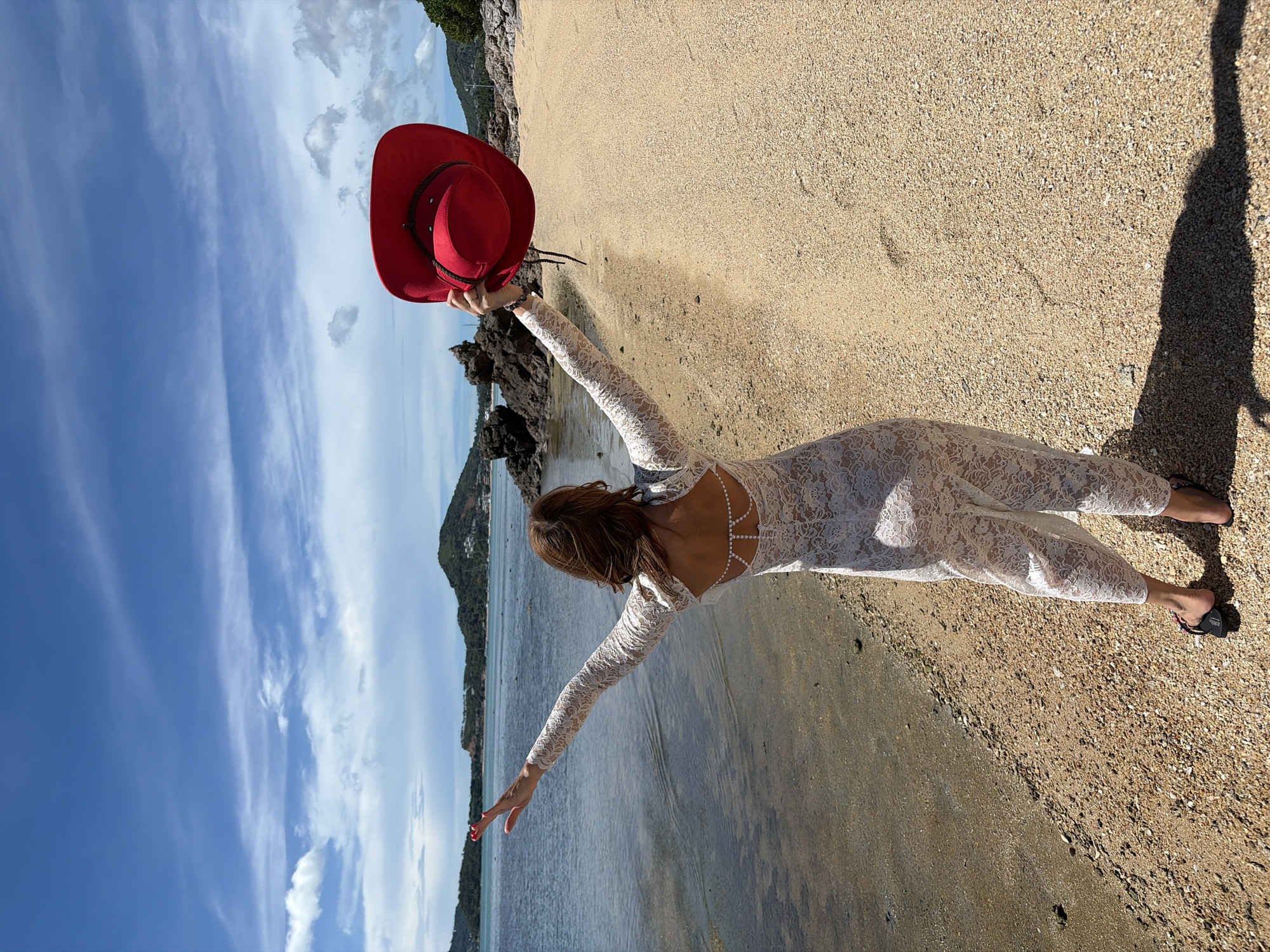 A person stands on a sandy beach in Baan Bo Phut, Thailand, facing away from the camera with arms outstretched. They are wearing a white lace jumpsuit and holding a red cowboy hat above their head in their right hand. The person is standing near the edge of the water, with the sea stretching out to the horizon. Hills covered in vegetation are visible in the distance, under a blue sky with scattered white clouds. The beach is composed of small pebbles and sand, and a shadow of the person is cast on the ground.