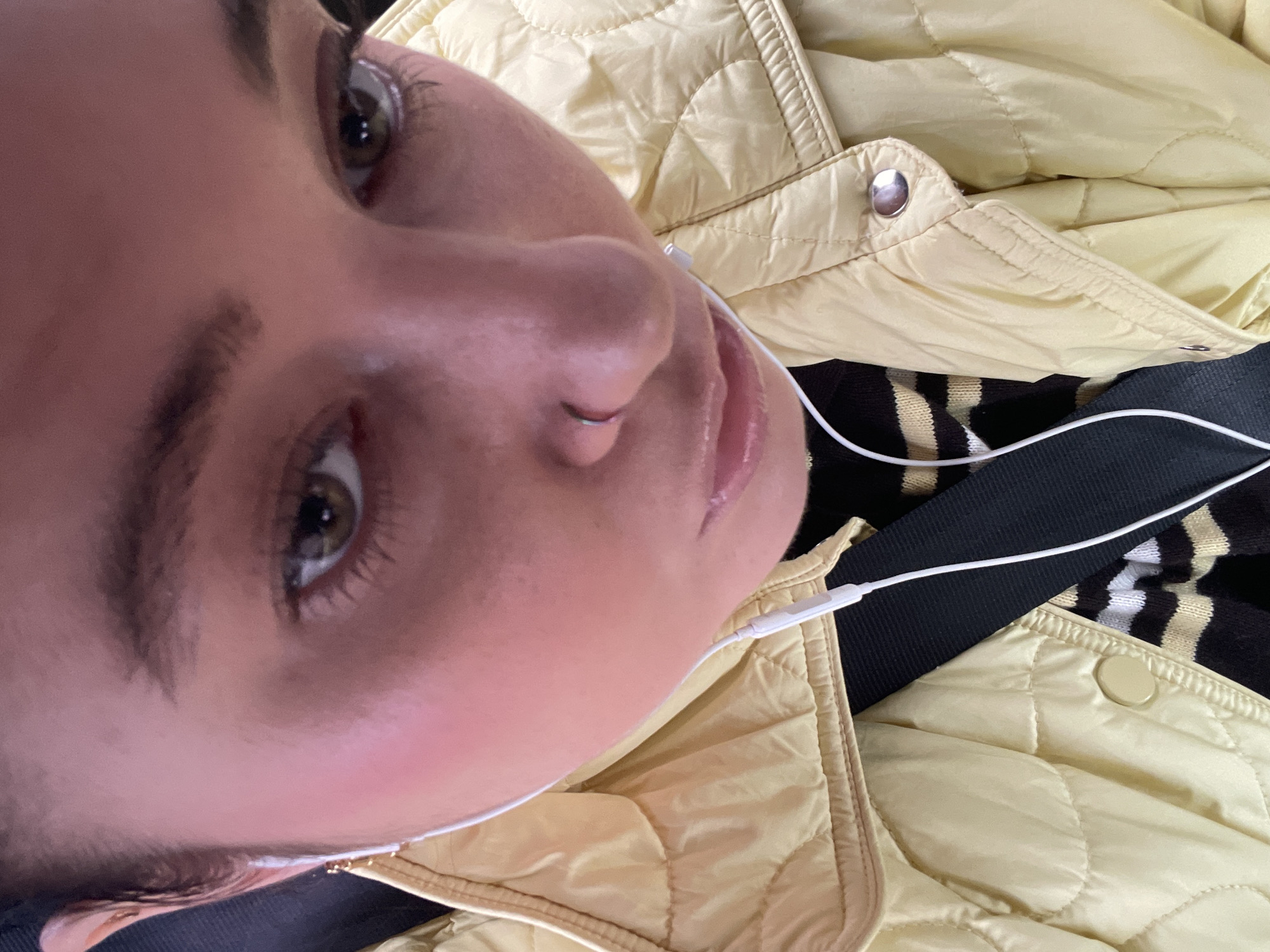 A close-up, slightly angled selfie-style image shows a person with light skin and brown eyes wearing a yellow quilted jacket. White earphones are in their ears. A black and white striped garment is visible beneath the jacket, along with a dark, possibly black, strap or fabric. A silver-colored nose ring is present in the person's left nostril. The background details are not discernible enough to specify the location as Moscow, Russia.