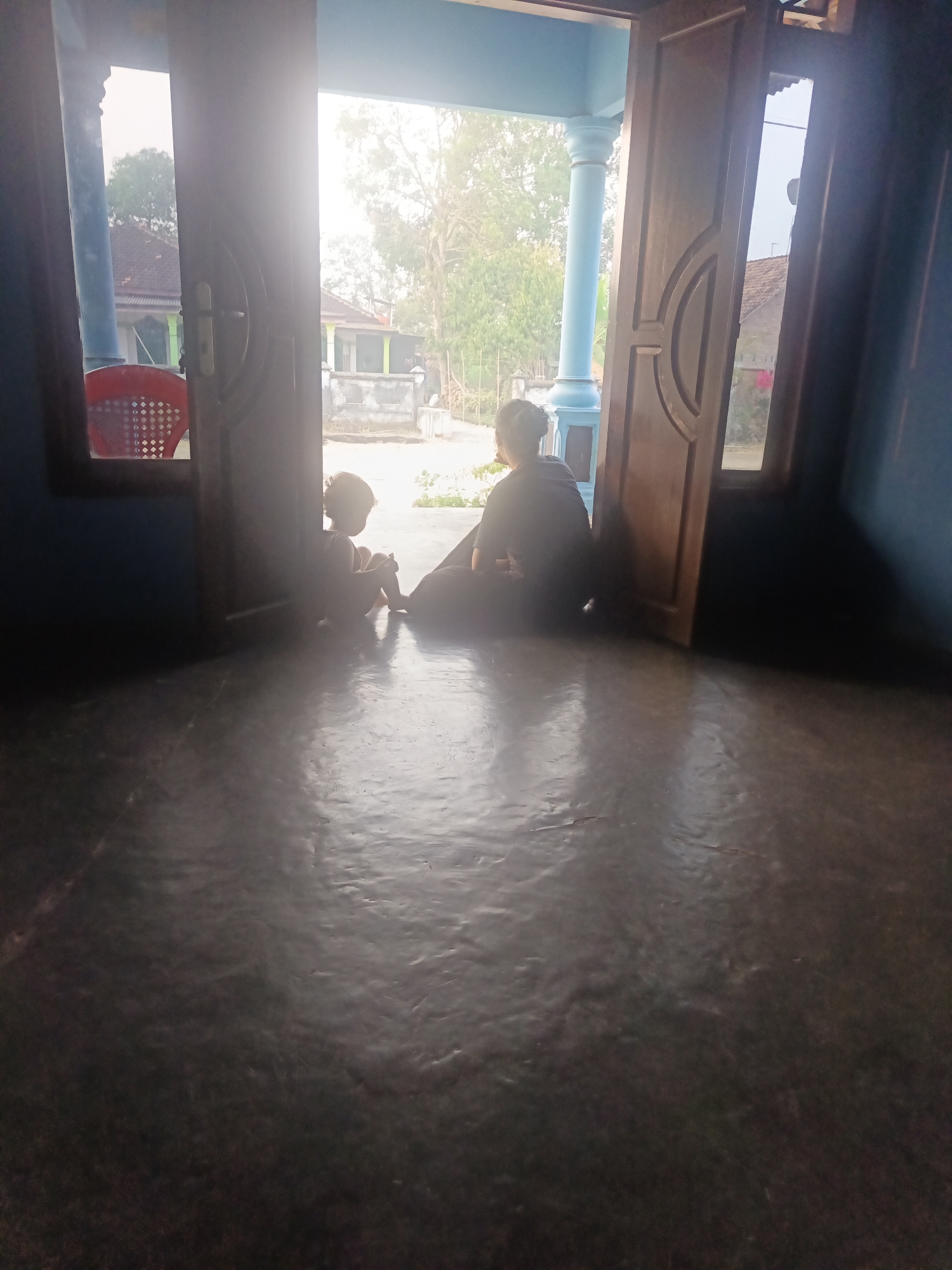 Two figures are silhouetted against a bright doorway opening into an outdoor scene. The figures are positioned on a reflective, dark gray floor inside a building, possibly a home. The figure on the left is smaller, appearing to be a child, sitting with their knees pulled towards their chest and looking outwards. The figure on the right is larger, likely an adult, also sitting and facing away from the camera, towards the doorway. The doorway reveals a house with a red plastic chair visible through a side window on the left, and trees and other buildings in the background. The scene appears to be in the city of Terbanggi Marga, Indonesia, suggested by the architectural style and vegetation. The overall lighting suggests it is daytime, with the bright light from outside creating strong backlighting and obscuring details of the figures and immediate surroundings.