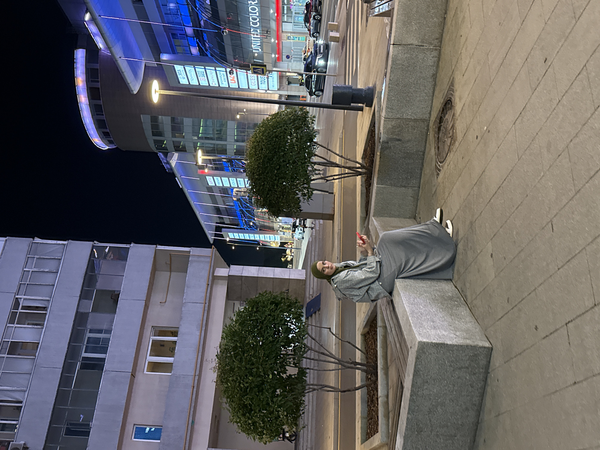 A woman wearing a hijab and a grey dress is sitting on a stone bench at night in front of a small tree. She is looking down at a red smartphone in her hands. Behind her, a modern building with many windows is illuminated by blue and white lights. The building appears to be a shopping center, with signs for various stores visible. Cars are driving on the street in front of the building, and streetlights illuminate the scene. The overall impression is of an urban environment at night. This scene is located at the intersection of Komsomolskaya Square and Kalanchevskaya Street in Moscow, Russia.
