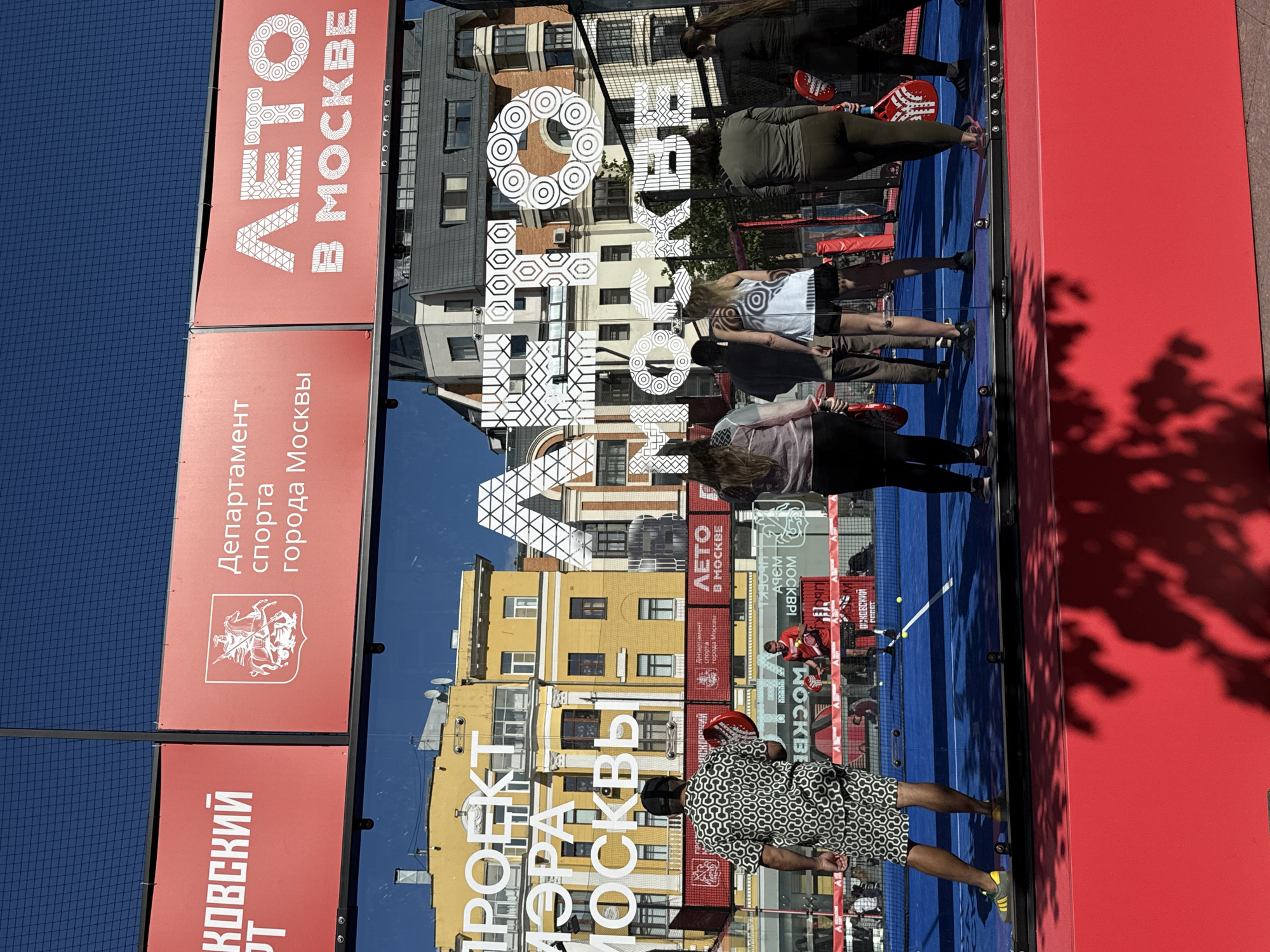 The image captures a vibrant outdoor scene, likely in Moscow, Russia, during what appears to be a sunny day, given the strong shadows. The primary focus is a large, red rectangular structure, possibly a wall or a large advertisement board, on the right side of the frame. This structure reflects the surrounding environment, creating a distorted but recognizable mirror image.
On the left side of the image, there are several red panels with white text and graphics. One panel features the text "Департамент спорта города Москвы" (Department of Sports of the City of Moscow) along with a white emblem depicting a knight on horseback. Another panel has "КОВСКИЙ" (Kovs'ky) written vertically in large white letters.
The reflection on the red surface reveals a scene with people engaged in an activity on what looks like a blue sports court, possibly for padel tennis given the presence of paddles. People are visible in athletic wear, with some holding red paddles. The reflection also shows buildings in the background, indicative of an urban setting, with warm-toned facades. Prominent text in the reflection includes "ЛЕТО В МОСКВЕ" (Summer in Moscow) and "ПРОЕКТ МЭРА МОСКВЫ" (Project of the Mayor of Moscow). The overall impression is one of a public space dedicated to sports and recreation, with a strong emphasis on a summer event or program in Moscow. The image is oriented vertically, as if the camera was held sideways.