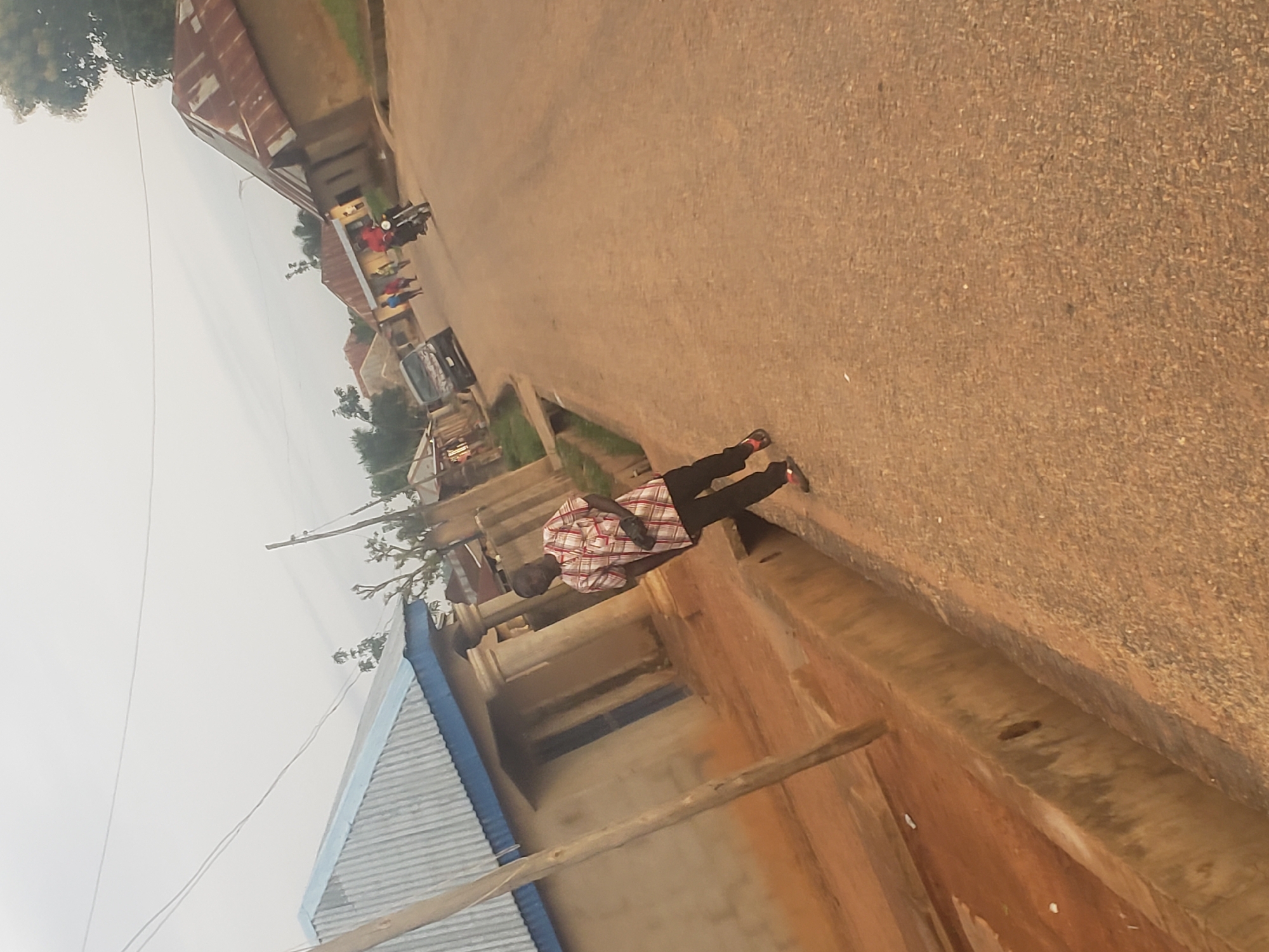 This is a street-level photograph taken in the city of Gumel, Nigeria. The scene depicts a street with buildings on either side and a few people and vehicles present. In the foreground, on the left, is a man wearing a checkered shirt and dark pants. He is standing on the edge of a drainage channel. To the right of the street, there are several buildings with corrugated metal roofs, typical of residential structures in the region. In the background, several other people can be seen walking along the street, and a motorcycle is visible. The overall environment appears to be a typical rural or semi-urban setting in Nigeria, with a dusty road and modest buildings. The sky is overcast, suggesting a cloudy day. The photograph has been taken at an angle, giving it a dynamic perspective.