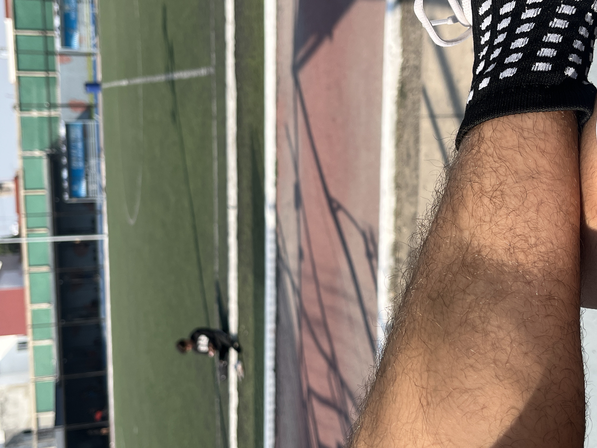 The image shows a person's leg wearing a black and white sock and white athletic shoes. The person appears to be elevated, overlooking a soccer field and a red running track. A person wearing black and white is on the soccer field. A building can be seen in the background. The location is Unknown, Unknown.