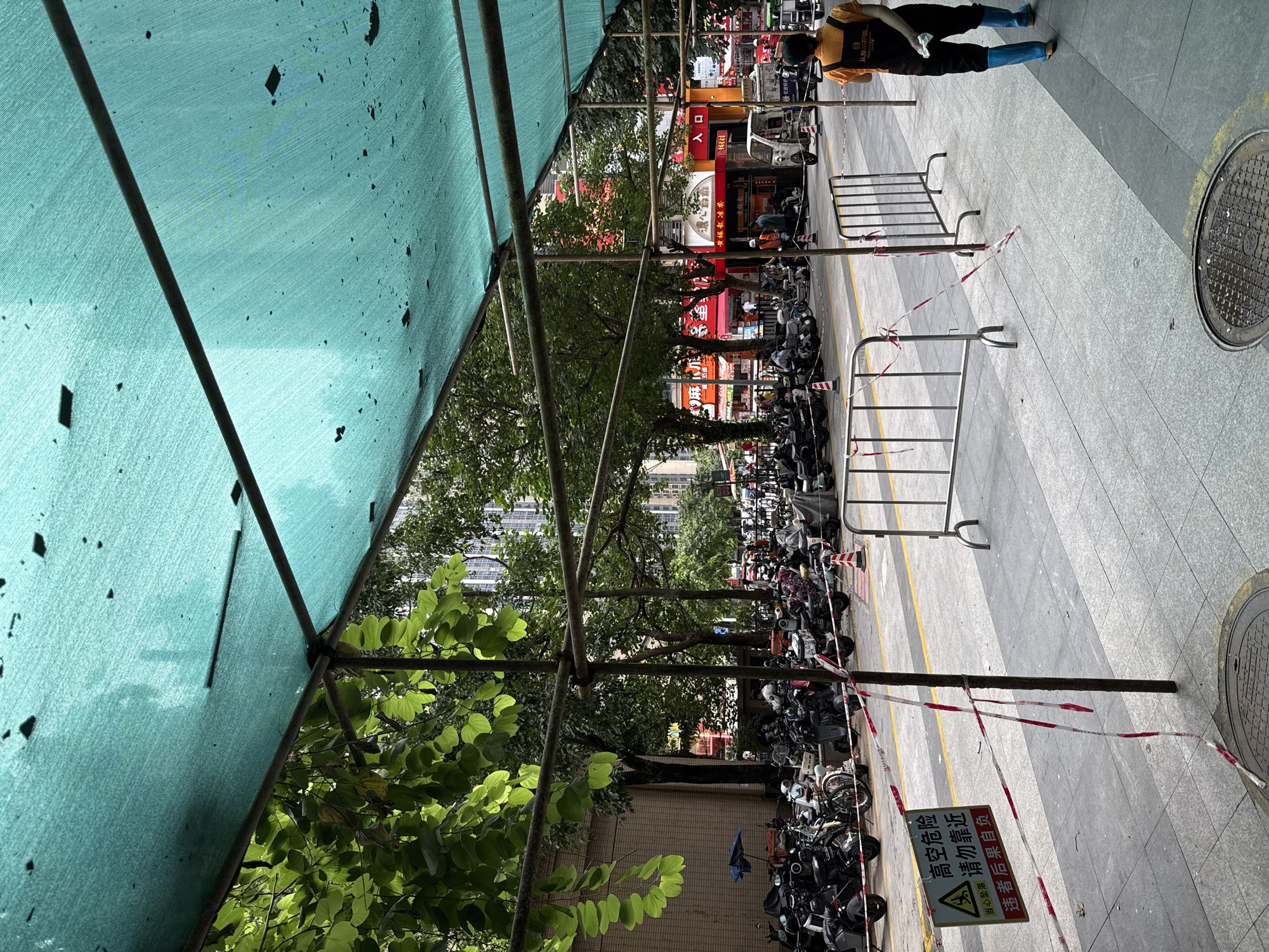 The image depicts a street scene in Haizhu District, China, characterized by a temporary overhead green shade structure supported by scaffolding. A dense row of parked motorcycles and scooters occupies the street, some with covers. In the foreground, a sidewalk is delineated by a yellow line, with metal barriers and red and white caution tape restricting access. A sign with Chinese characters, warning of "high altitude danger" and advising against approaching, is visible. A person wearing an orange vest and dark pants, with blue boots, is walking away from the camera on the sidewalk. Trees with green foliage line the street, and buildings with visible windows are in the background. Several storefronts with red and yellow signage are also present. The overall impression is one of urban activity with temporary safety measures in place.