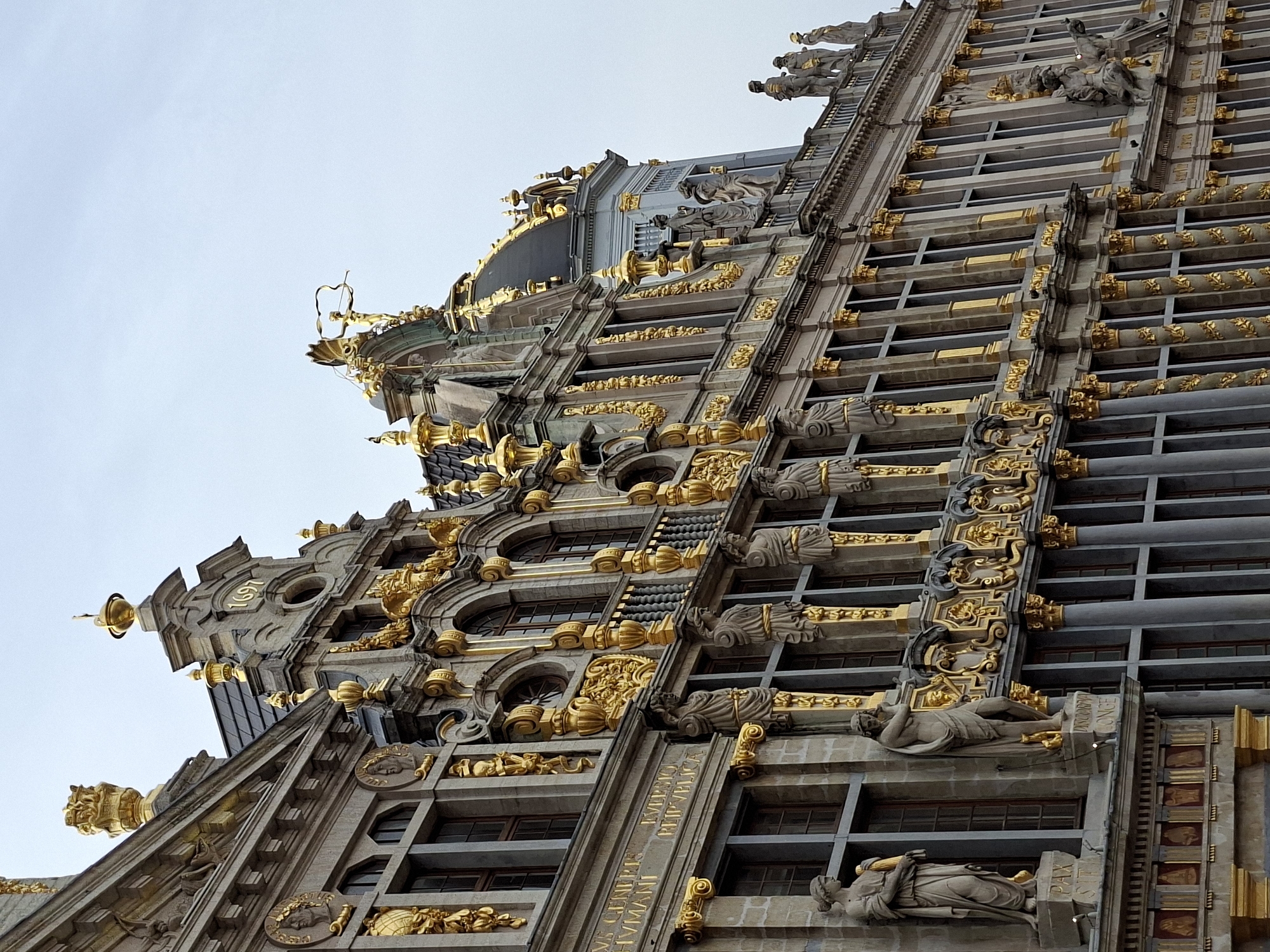 The image captures the ornate facade of a historic building in Brussels, Belgium. The architecture is characterized by its Baroque style, featuring extensive gold ornamentation, including statues, columns, and decorative elements. The building's stone facade is intricately detailed, with multiple windows and balconies. A clear sky forms the background. The specific location in Brussels is the Grand Place, one of the most famous public squares in the city. The building appears to be one of the Guildhalls that line the square. The detail of the stonework and the gold accents suggest it is a building of significant historical and architectural importance.
