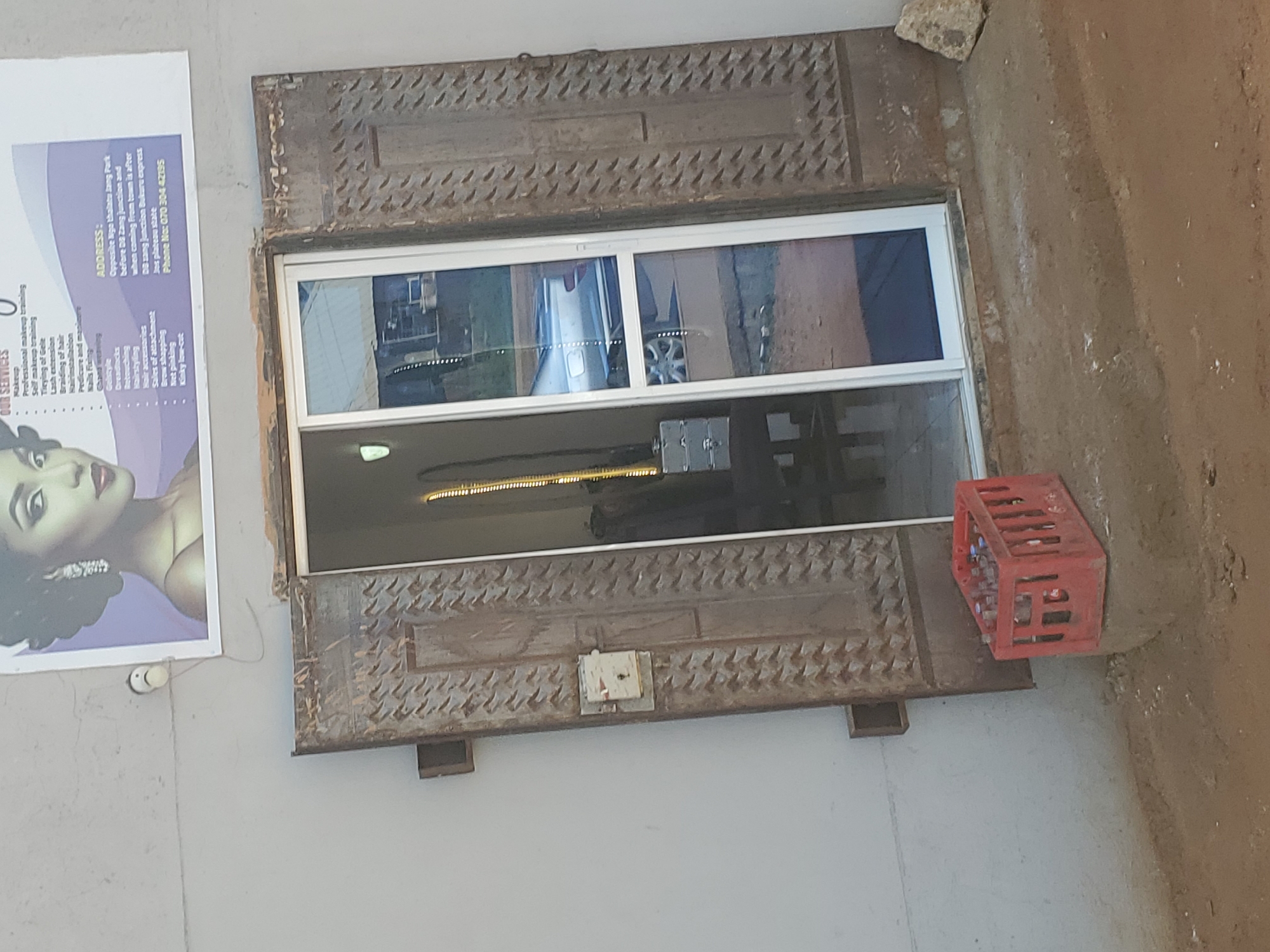 A medium shot shows the entrance to a business in Bukuru, Nigeria. The entrance is framed by heavy, decorative metal doors that are partially ajar, revealing a glass sliding door behind them. To the left of the entrance, a red plastic crate is filled with glass bottles, some of which appear to be sodas. Above the entrance, a poster displays an image of a woman and lists business services and an address in Bukuru, Nigeria, along with a phone number. The business appears to be a salon or beauty shop. The ground in front of the entrance is covered in dirt and gravel, suggesting an outdoor area. The wall behind the poster is a light gray, and the overall scene has a somewhat dilapidated feel.