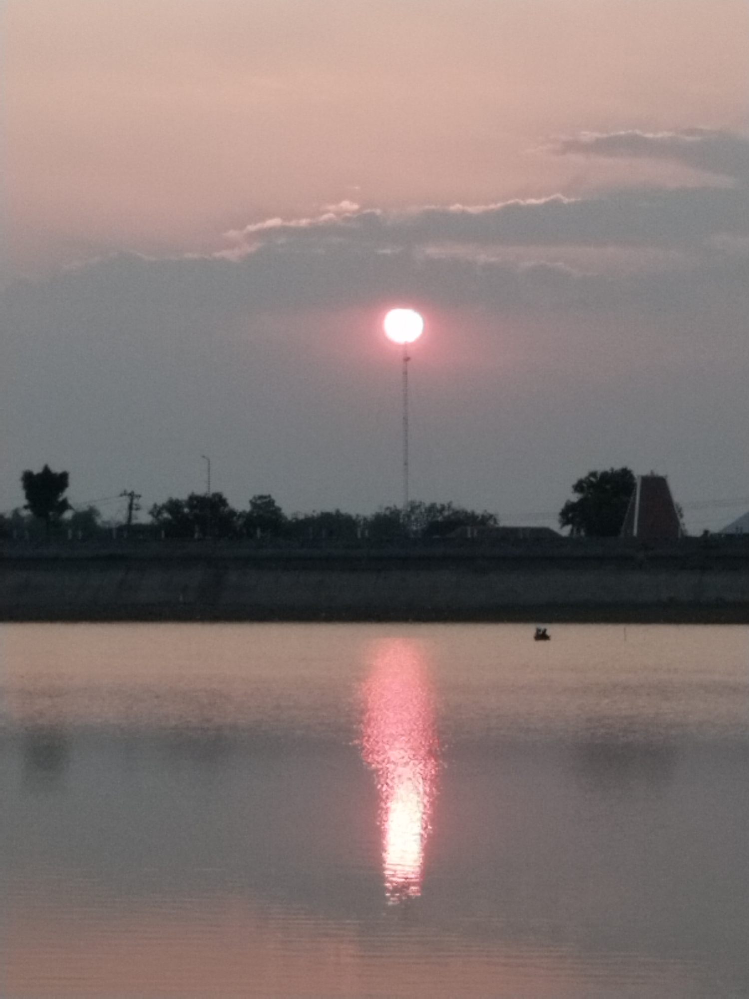 The image depicts a sunset over a body of water, likely a reservoir or a lake, in Sambirejo, Indonesia. The sun, a bright pinkish-red orb, is partially obscured by thin clouds in the upper center of the frame. A reflection of the sun streaks down the water's surface, creating a shimmering path of light. Along the far bank, silhouetted against the dusky sky, are trees and structures. One prominent structure is a tall, slender tower topped by a small circular object, likely a communication tower. To the right, a building with a distinctive pyramid-shaped roof, possibly a temple or a unique architectural feature, is visible. In the middle distance on the water, a small dark object, appearing to be a boat with two occupants, is present. The overall atmosphere is tranquil and serene, characteristic of a sunset.