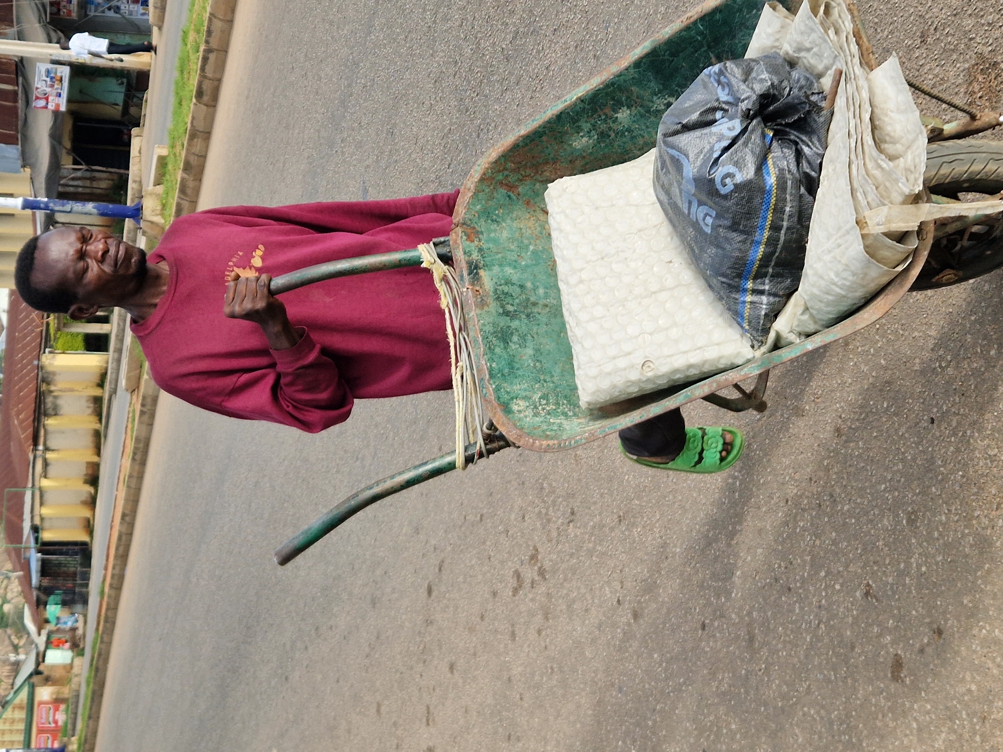 A dark-skinned man with a weary expression pushes a green, rusty wheelbarrow down a paved road in Kabong, Nigeria. He is wearing a maroon sweatshirt and bright green flip-flops. The wheelbarrow contains a white, textured mat and a black plastic bag with blue and yellow stripes. In the background, there are shops and buildings lining the street, some with corrugated metal roofs. A man in a white shirt is visible further down the road. The sky is overcast.