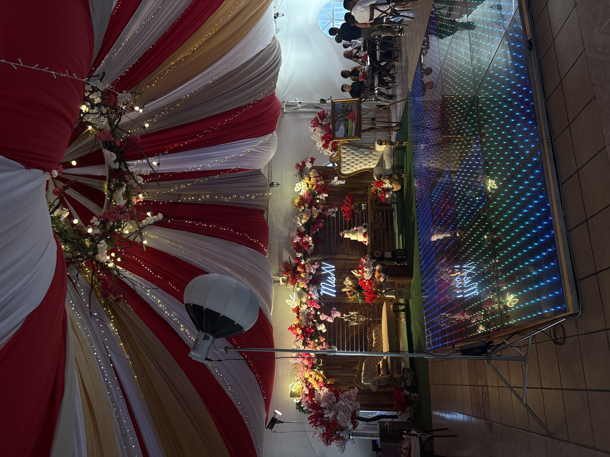 The image shows an event space in Chilpancingo, Mexico, likely for a quinceañera. The space is decorated with red, white, and gold draped fabric with string lights. There is a backdrop with the words "Mis XV" in neon lighting, surrounded by floral arrangements. A white chair is positioned in front of the backdrop. A lighted dance floor is present, and several people are seated at a table in the background. An Amaran 300c lighting unit is visible.