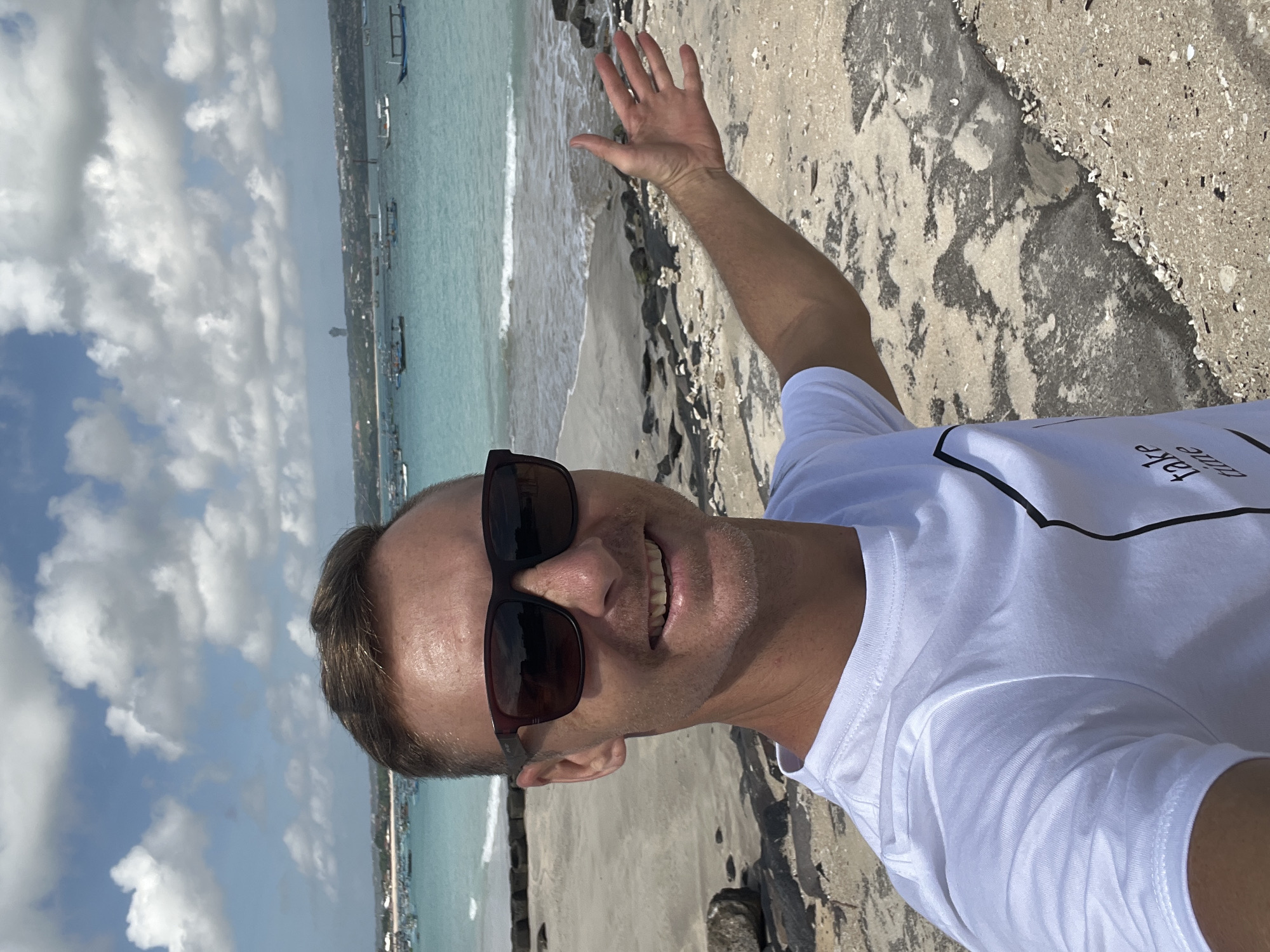 A man in a white t-shirt and sunglasses smiles at the camera, holding his right arm out with his palm facing upwards and fingers spread. He is standing on a sandy beach with scattered rocks and a concrete pathway. In the background, the turquoise water of the ocean is visible, dotted with numerous small boats. The sky is partly cloudy, with a line of buildings and a tall structure visible in the distance. The scene is likely in Kuta, Indonesia, a popular tourist destination known for its beaches and vibrant atmosphere.