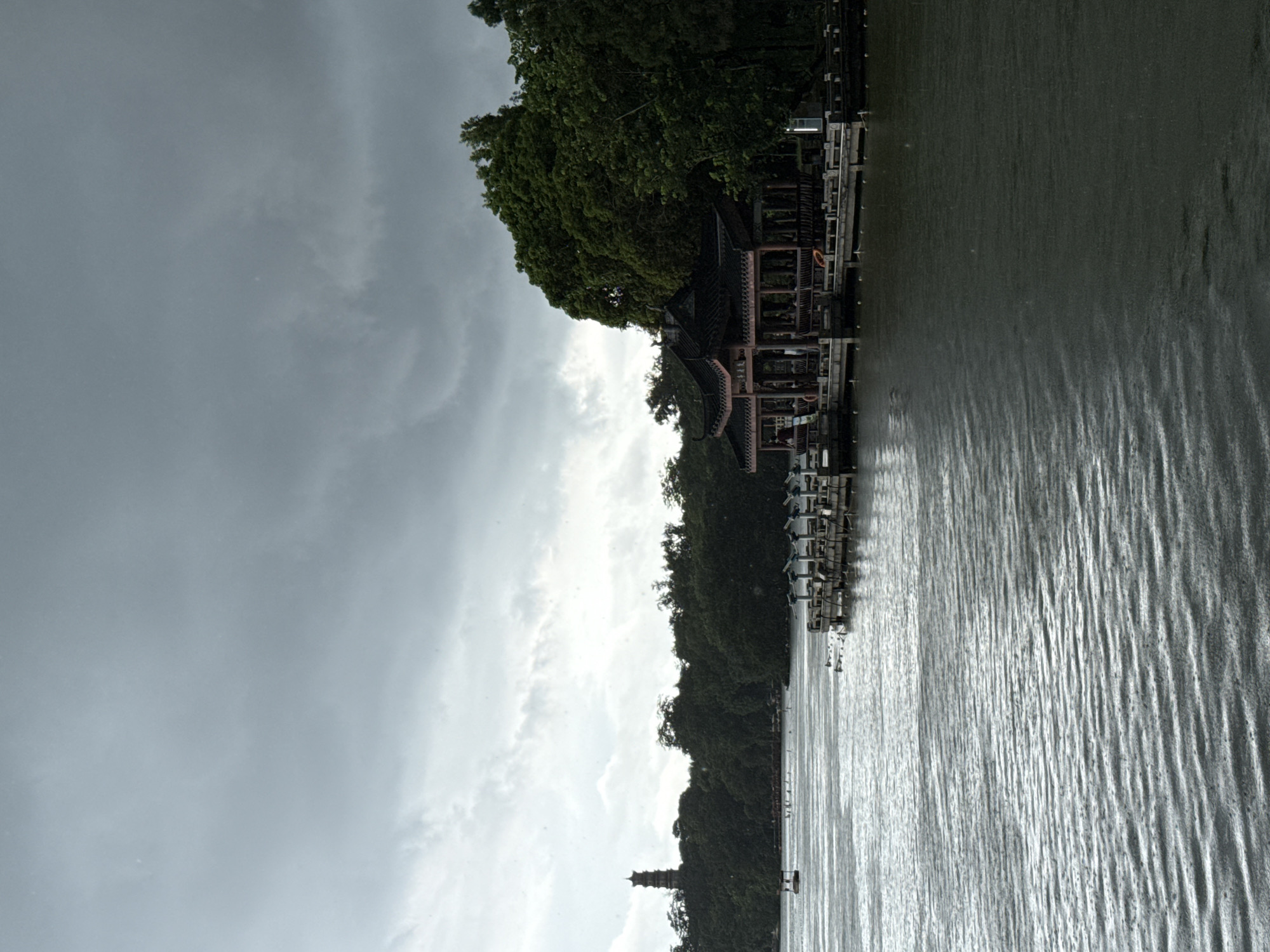 This is a medium shot, eye-level, horizontal, full-color, high-angle photograph of a lake with a pavilion and a pagoda on its shore, set against a dark, overcast sky. The scene is likely the West Lake in Huizhou, China, given the distinctive pagoda and the architectural style of the pavilion.
In the foreground, the dark, rippling surface of the lake dominates the lower half of the image. The water has a metallic sheen due to the overcast light. Small white dots, indicative of rain, are visible on the water's surface and in the air.
To the right, a wooden pavilion with a dark, multi-tiered, curved roof stands on a wooden pier extending into the lake. The pavilion is open-sided, revealing rows of dark wooden benches and several figures, some appearing to be patrons or staff. There are safety buoys visible on the pier.
In the background, a dense line of dark green trees forms the shoreline. On the far left, a tall, multi-story pagoda with a pointed roof rises above the treeline. A small, light-colored boat is visible on the lake in the middle ground, with other indistinct figures or objects on the water further in the distance.
The sky is a turbulent mass of dark grey and white clouds, suggesting imminent or ongoing rain. The overall mood of the image is somber and atmospheric, enhanced by the weather conditions and the architectural elements.