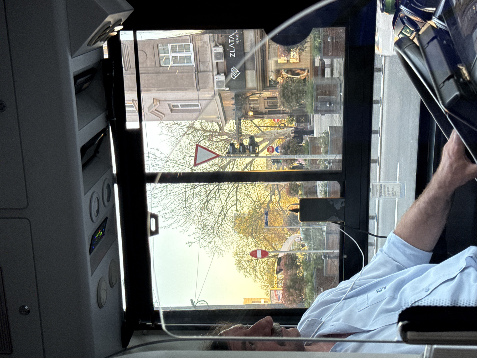 The image shows a view from inside a vehicle, possibly a bus or train, looking out the window. A man in a light blue shirt is partially visible in the lower right and bottom of the frame. Outside the window, a street scene unfolds, with buildings, trees, and traffic signs visible. A building with the name "ZLATA NAKIT" is recognizable. There is a triangular traffic sign and a red and white round sign, likely indicating a prohibited entry. The light suggests it might be late afternoon or early evening. The location is likely Belgrade, Serbia, based on the name "ZLATA NAKIT".