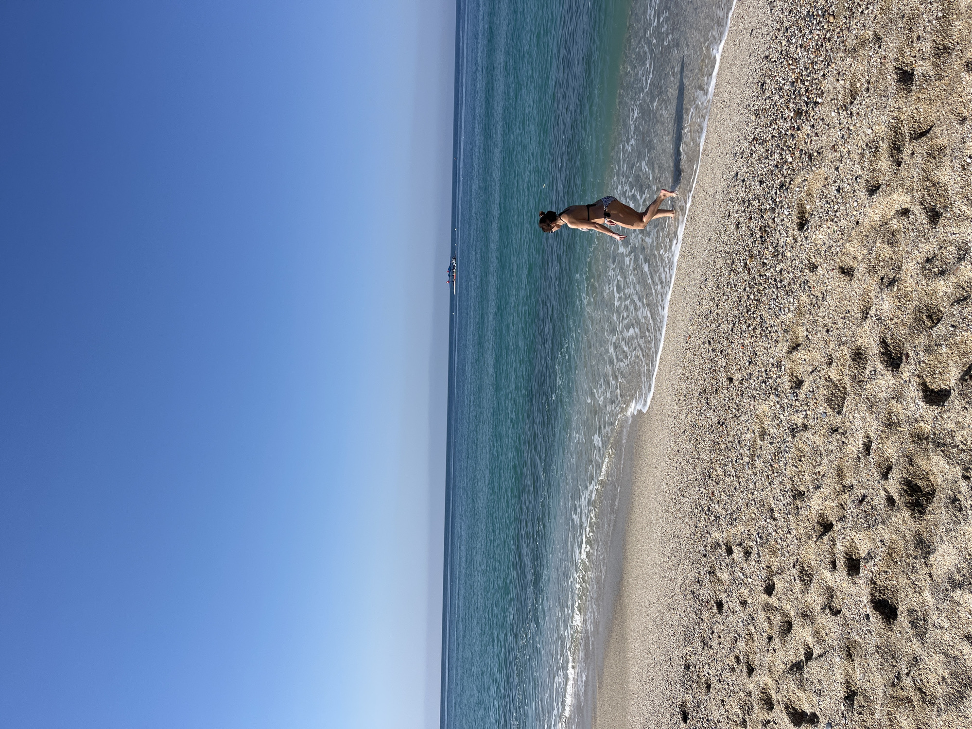 This image depicts a sunny day on a beach, likely in Alanya, Turkey, with a clear blue sky meeting the horizon over the sea. A person, identified as a woman wearing a bikini, is seen in the shallow, clear blue-green water at the edge of the shore. She appears to be in an active pose, leaning forward with her arms extended towards the water, almost parallel to the surface, as if dipping her upper body into the sea or performing a playful dive/stretch. Her feet are in the foamy part of a gentle wave.
The beach consists of a mix of light-colored sand and small pebbles, with visible footprints scattered across the sandy areas. Gentle waves are breaking on the shore, creating a line of white foam. The vast expanse of the sea stretches out to the horizon, and in the far distance, a small boat or ship can be faintly seen. The sky above is a uniform, clear blue, indicating bright, sunny weather. No text is visible in the image.