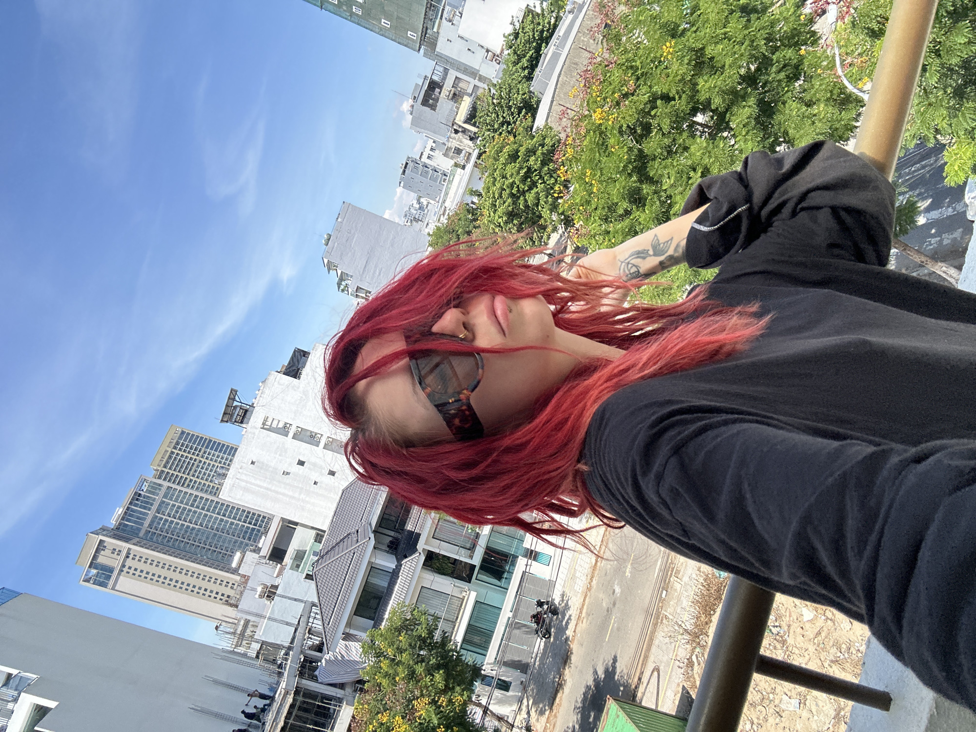 A woman with bright red hair and sunglasses is taking a selfie on a balcony overlooking a city. The city appears to be Đà Nẵng, Vietnam, with several tall buildings and trees visible in the background. The woman is wearing a black long-sleeved shirt. The balcony railing is in the foreground. There are some construction activities and a motorbike on the street below. The sky is blue with a few clouds.