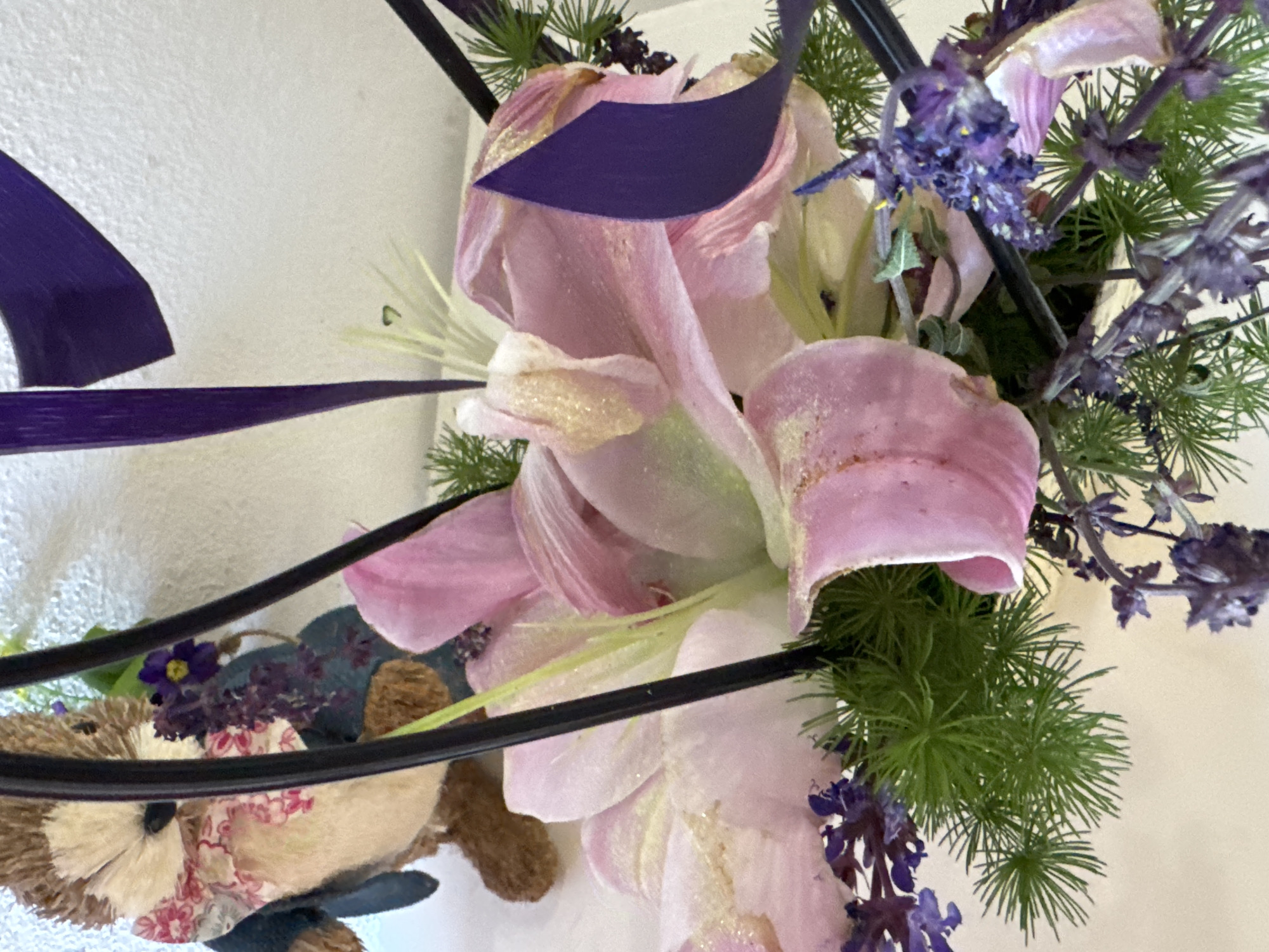 The image depicts an arrangement of flowers with a toy hedgehog incorporated into the design. The flowers are primarily lilies in various shades of pink and white, accented with purple flowers and green foliage. Dark purple ribbons are woven through the arrangement. The hedgehog toy is wearing a patterned dress and blue vest. The background is a plain white wall. No information about Sofia, Bulgaria is present in the image.