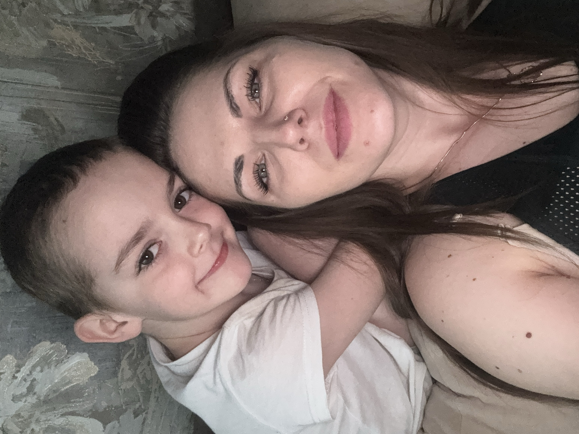 The image shows a woman and a young child lying down together. The woman has brown hair, fair skin, and a nose piercing. She is wearing a black top and a necklace. The child is wearing a white t-shirt and has short, dark hair. They are both smiling slightly and looking at the camera. The background appears to be a patterned couch or wall. The location cannot be determined based on the image content.