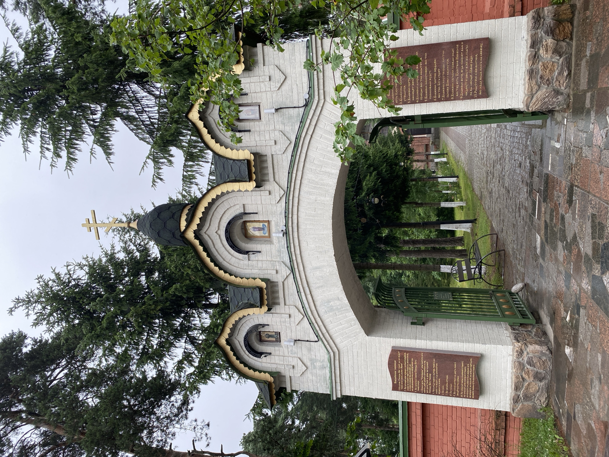 The image shows the ornate entrance gate to a religious complex, likely a monastery or church, in Priozersk, Russia. The gate is constructed from white brick with arched openings and decorative gold trim. A central, green-domed structure topped with a golden Orthodox cross stands above the gate. Three arched niches are visible on the gate's facade, each containing a religious icon. The path leading through the gate is paved with cobblestones and lined with lush green trees. A red brick wall flanks the left side of the entrance. Two large plaques with text in Cyrillic script are mounted on the gate's pillars. The overall atmosphere is serene and reflects a historical and spiritual site.