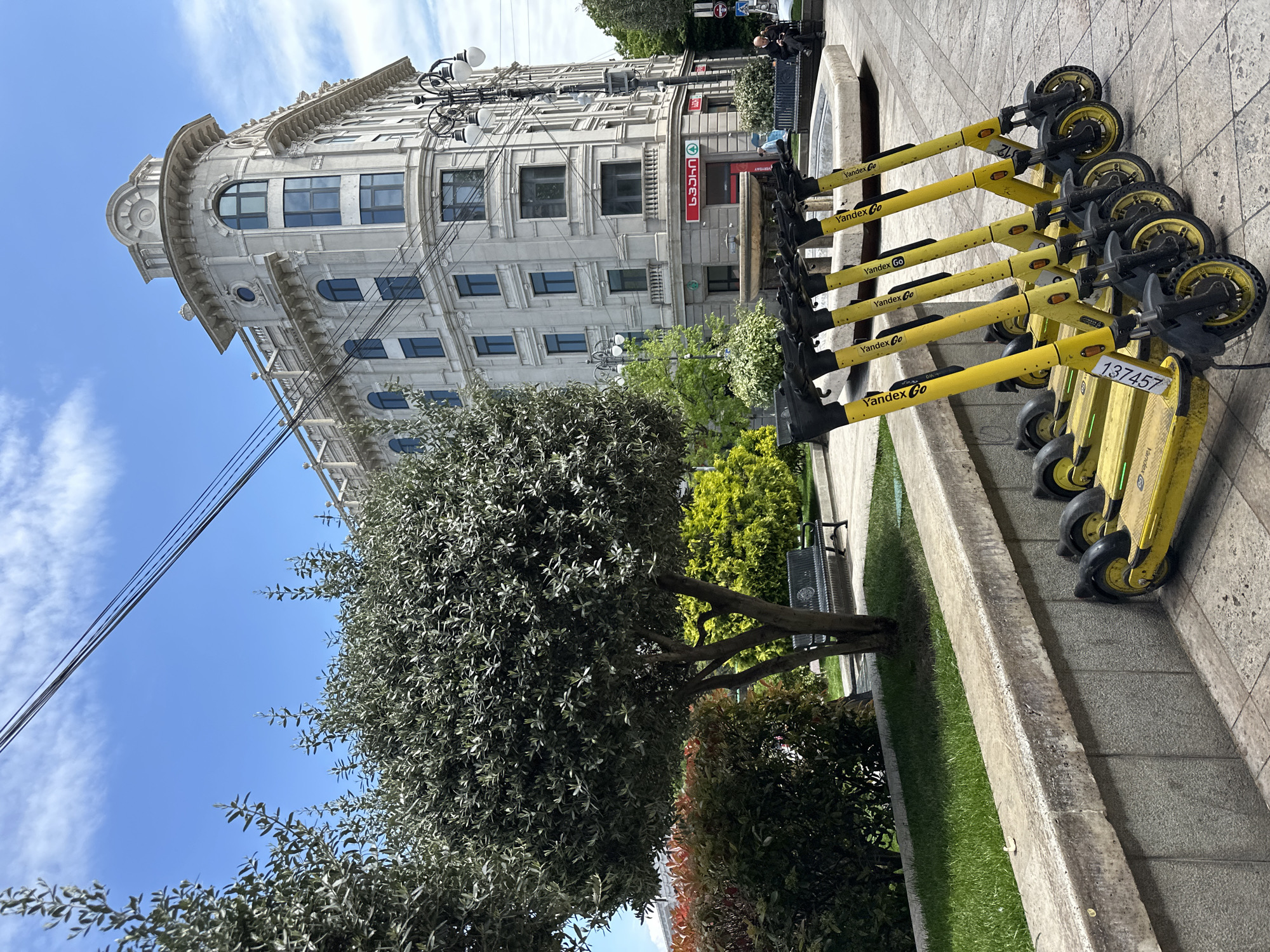 This media file depicts an urban street scene in Tbilisi, Georgia, under a bright, clear blue sky with scattered wispy clouds, indicating daytime.
In the foreground, a row of yellow electric scooters, branded "Yandex Go," is neatly parked on a grey tiled sidewalk. One scooter displays the identification number "137457." The sidewalk is bordered by a concrete curb, beyond which lies a strip of green grass and various trees and bushes, including a large, full-leaved tree. A metal park bench is partially visible among the greenery.
The background is dominated by a grand, multi-story building with a light-colored facade, featuring ornate architectural details, numerous windows (some arched, some rectangular), and a distinctive rounded corner with decorative elements at its top. On the ground floor of this building, a shop entrance is marked by a red sign displaying Georgian text that reads "სუპერ" (Super) and "AYGEBA" (meaning "rise" or "development").
Overhead, several utility power lines stretch across the frame. Multiple ornate streetlights with globe fixtures are also visible along the street. In the distance, a couple of people can be seen near the building, suggesting a public area. The scene combines historic architecture with modern urban transport options.