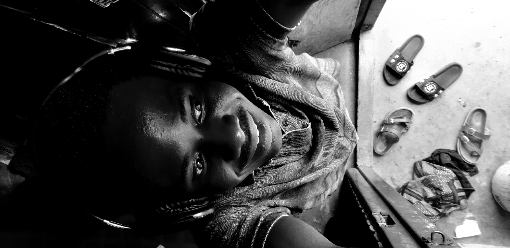 In a high-angle, black and white photograph taken in the city of Narayi, Nigeria, a young man wearing headphones is looking directly at the camera with a wide smile. He is wearing a collared shirt. Below him, on a concrete surface, are several pairs of sandals and a folded piece of fabric. The background is dark and indistinct, with a hint of a textured surface behind the man. The scene appears to be indoors, possibly in a room or outdoor shelter, with a simple, somewhat utilitarian environment.