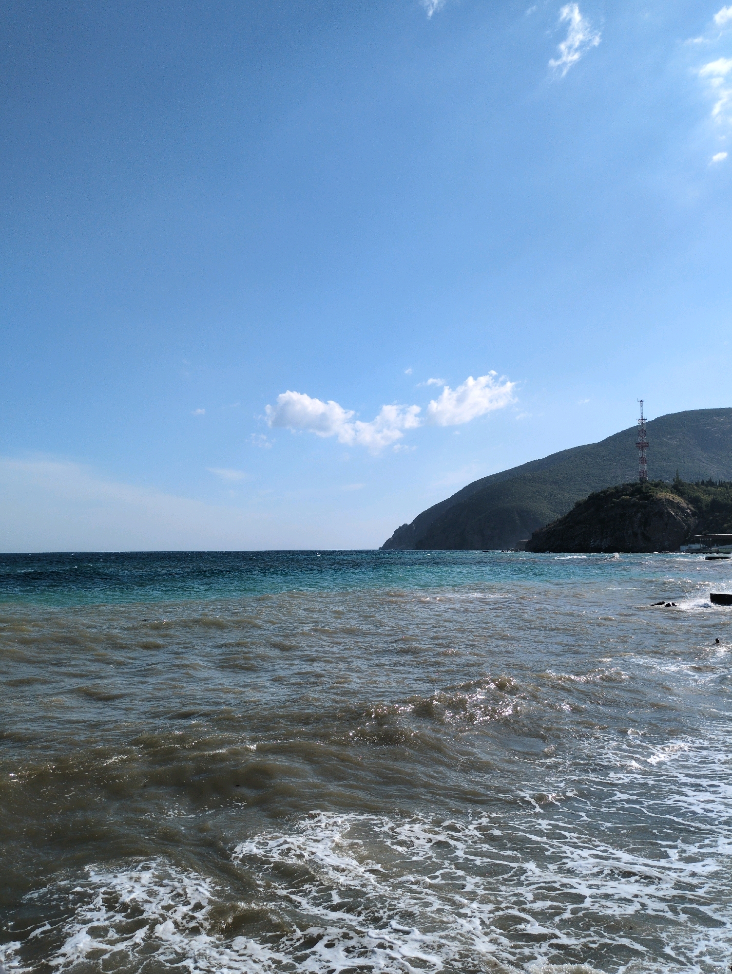 The image shows a coastal landscape in Ukraine. The scene is dominated by the sea and a hilly terrain. The sea is divided into two distinct bands of color: a darker, muddy brown closer to the shore, and a clearer, turquoise blue further out. Waves are breaking along the shore, creating white foam. In the distance, a large, green hill rises steeply from the coast. A red and white communication tower is visible on top of a smaller, rocky outcrop near the base of the hill. A few scattered white clouds are present in the clear blue sky. The overall impression is of a natural coastal environment with some man-made infrastructure.
