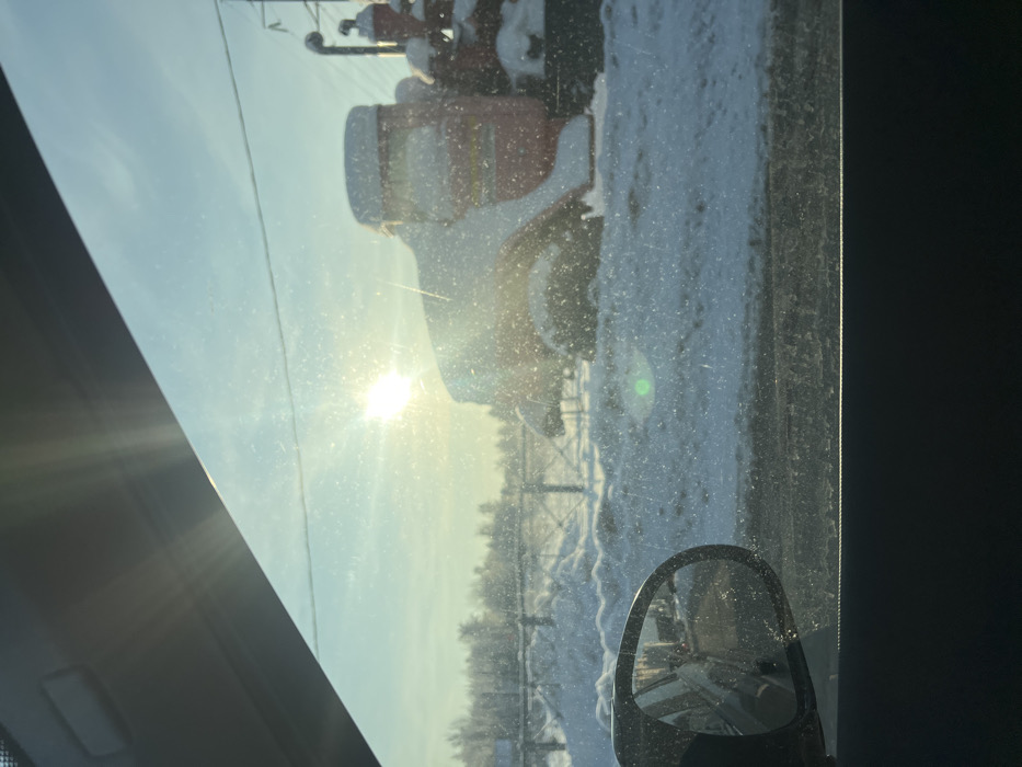 A winter scene in Кальтино, Russia, is observed from inside a vehicle through a dirty side window. The bright, low-angled sun, positioned slightly left of center, causes significant glare and lens flare across the frame. On the right, a large, stationary red industrial vehicle, likely a tanker truck, is heavily covered in white snow. The ground beneath the vehicle and extending into the middle ground is entirely covered in snow, with irregular drifts visible. A light-colored wooden fence with diagonal supports runs horizontally in the background, behind the snowdrifts. Further back, a line of bare deciduous trees stands against a light blue sky. The foreground includes the dark interior trim of the viewing vehicle, a section of the headliner in the upper left, and a black side mirror in the lower left reflecting a blurred street environment. No individuals or active elements are present.
