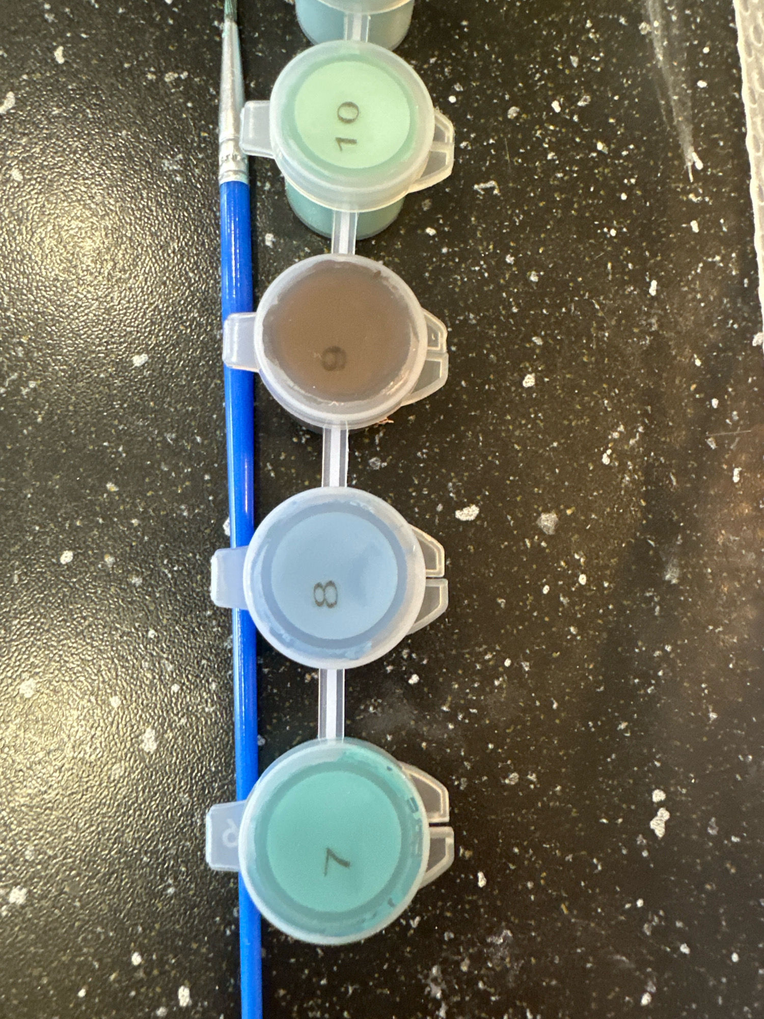 The image shows a top-down view of several small, round paint pots, each with a clear plastic lid. A blue-handled paintbrush with silver bristles is positioned vertically to the left of the paint pots. The paint pots are lined up in a row and appear to be connected by thin plastic strips. Each pot contains a different color of paint. From top to bottom, the visible numbers on the lids are "10", "9", "8", and "7". The paint in pot "10" is light green, pot "9" is brown, pot "8" is light blue, and pot "7" is also light green. The surface beneath the paint pots and brush is a dark, speckled material, possibly a countertop. There is no indication of the location within the city of Nevinnomyssk, Russia, or any people, actions, or interactions depicted in the image.