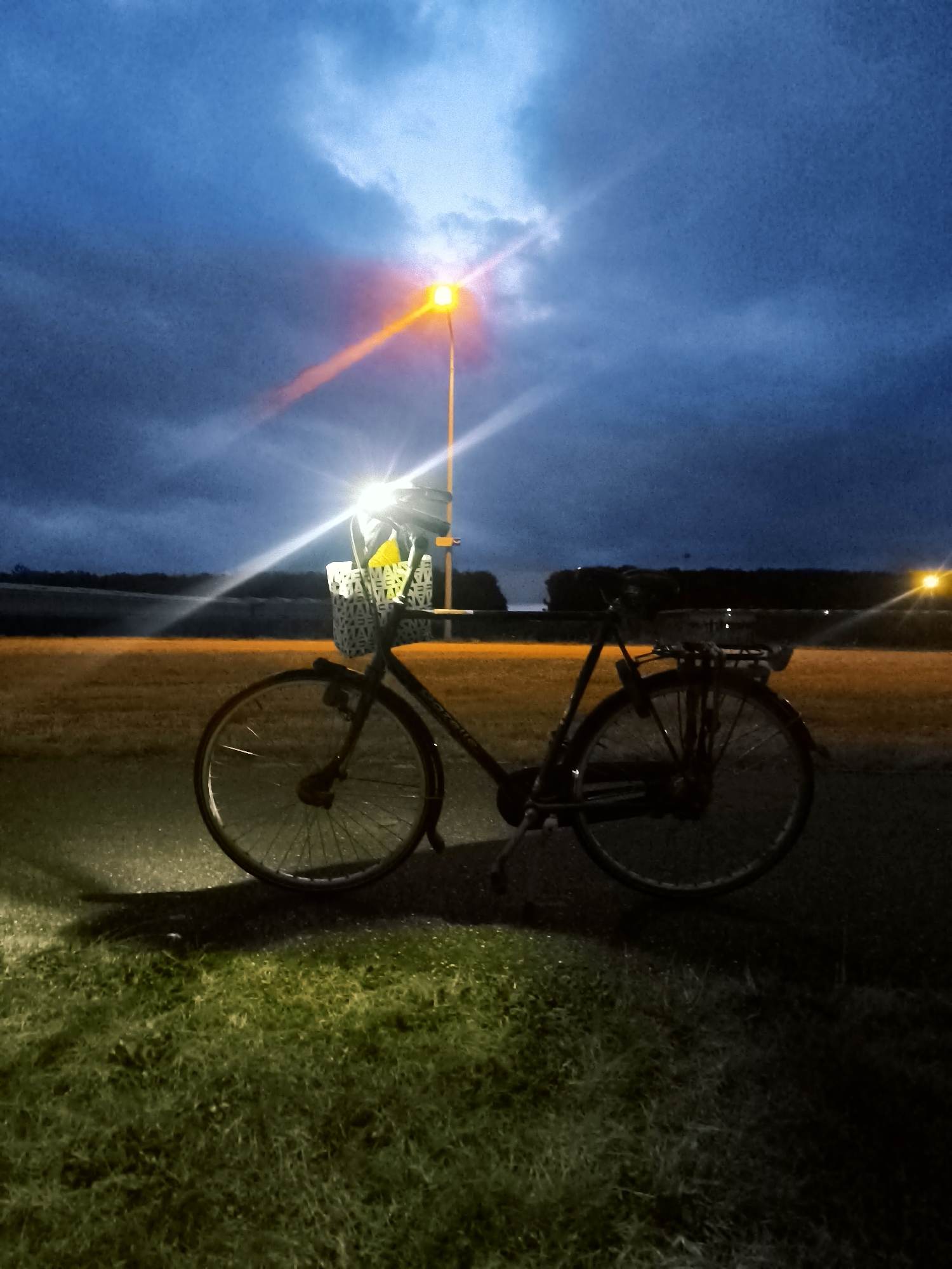 The image depicts a bicycle parked on an asphalt path at dusk or dawn, likely in Almere, Netherlands. A street lamp overhead illuminates the scene, casting a strong beam of light that reflects off the wet pavement and illuminates the bicycle's front wheel and basket. The bicycle is black with a white basket containing a yellow object. In the background, a row of trees forms a dark line against a twilight sky filled with dramatic clouds. Another street lamp is visible in the distance on the right side of the image. The ground in the foreground is covered in grass, which is also lit by the street lamp. The overall atmosphere is moody and slightly surreal due to the dramatic lighting and twilight conditions.