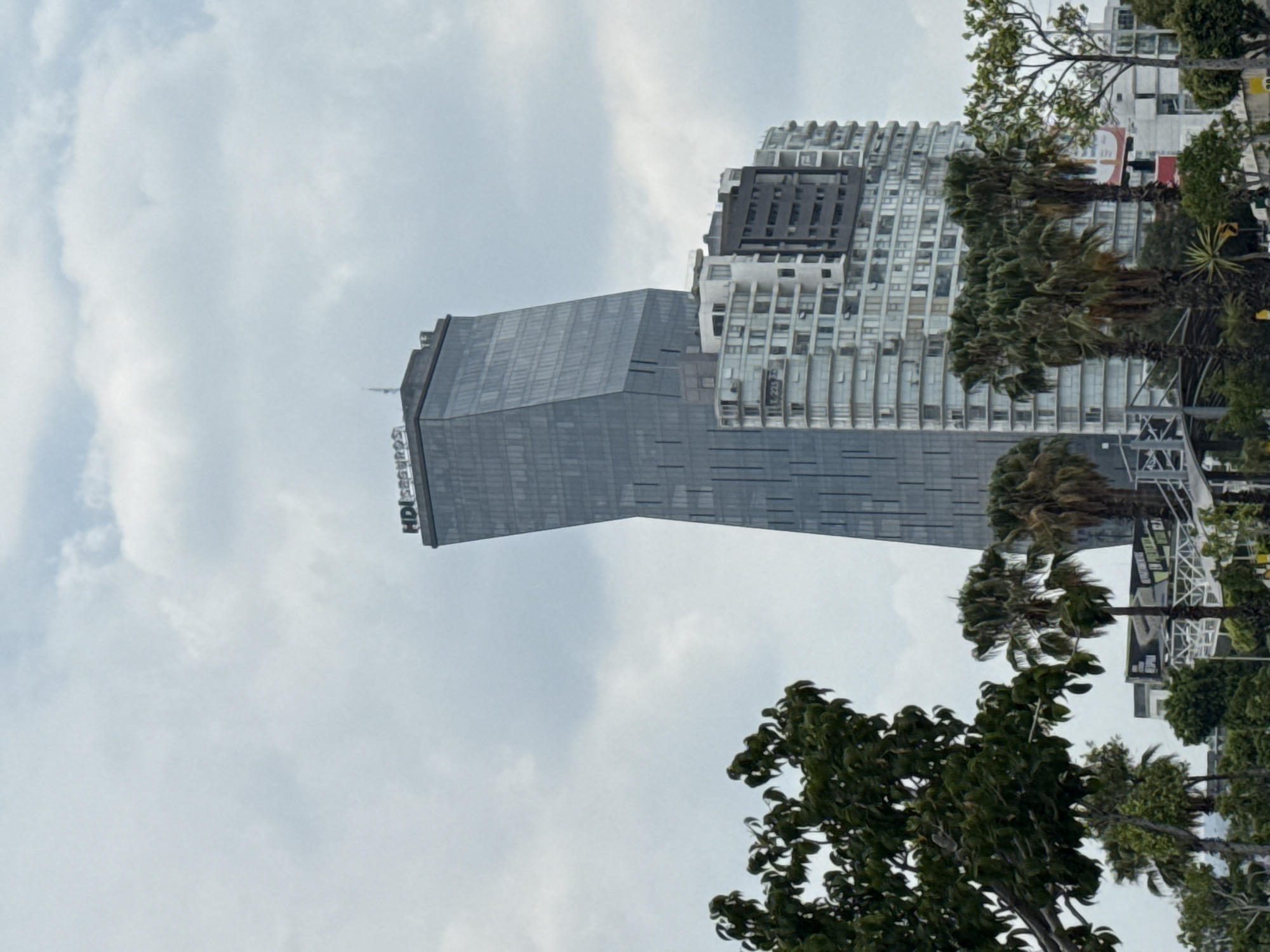 A medium shot, taken outdoors in Puebla, Mexico, depicts two tall buildings under a cloudy sky. The building on the left is a modern, geometric skyscraper with the "HDI Seguros" logo visible on its side. The building on the right is a high-rise residential building with many balconies. Trees are visible in the foreground.
