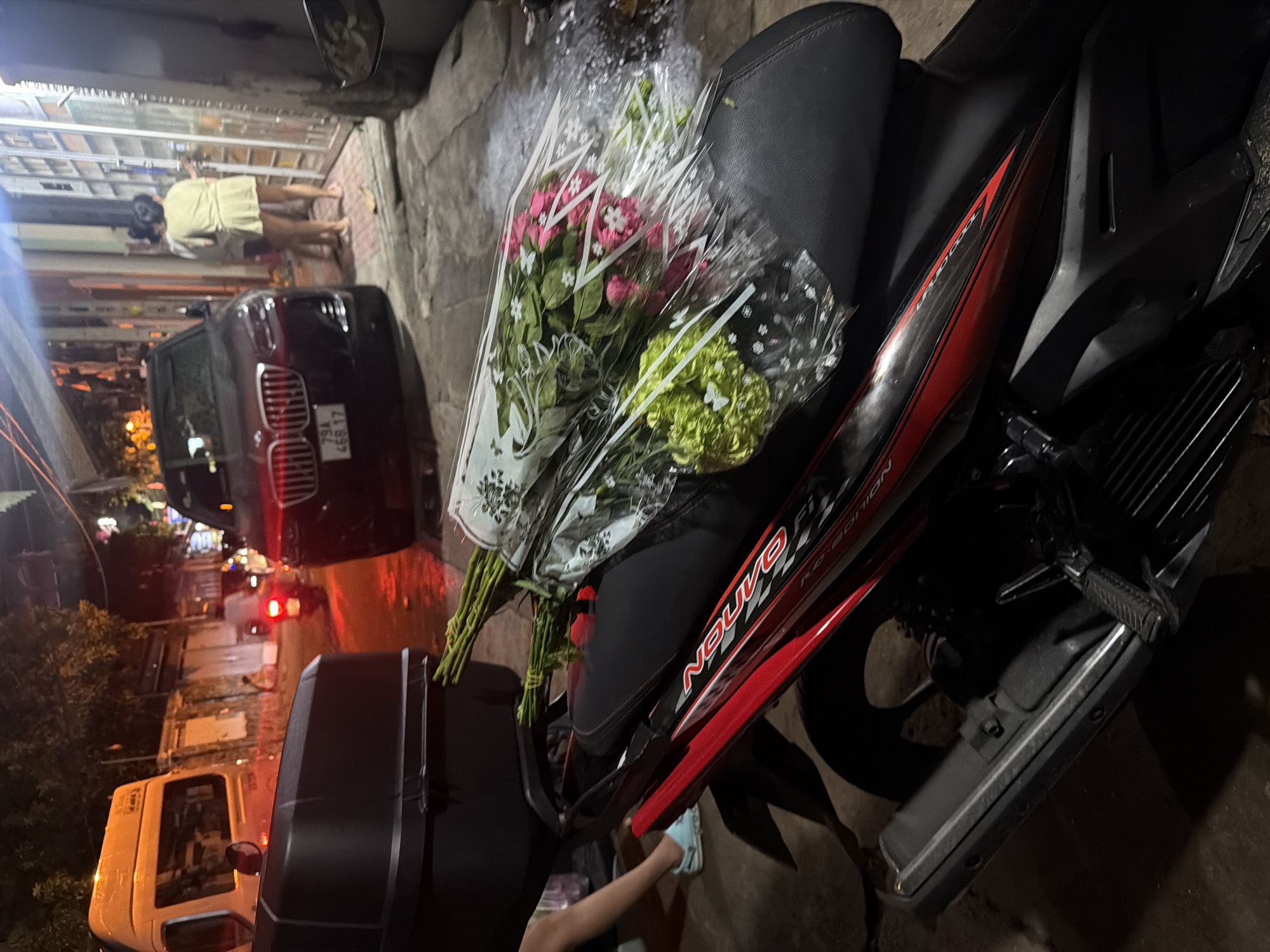 The image depicts a street scene in Nha Trang, Vietnam, at night. In the foreground, a black motorcycle is partially visible, with a large black storage box mounted on the back. Two bouquets of flowers, one with pink flowers and the other with green flowers, are placed on the seat of the motorcycle, wrapped in clear plastic. The motorcycle has a red and black design on its side.
In the background, a dark-colored SUV is parked on the street, with a license plate that reads "79A 468.17". Further down the street, a white van is parked. Two women are standing near a building entrance on the right side of the image, one of whom is wearing a light-colored dress. The street is illuminated by streetlights and the lights from buildings. The overall impression is a typical urban streetscape at night, with a focus on the motorcycle and the flowers.