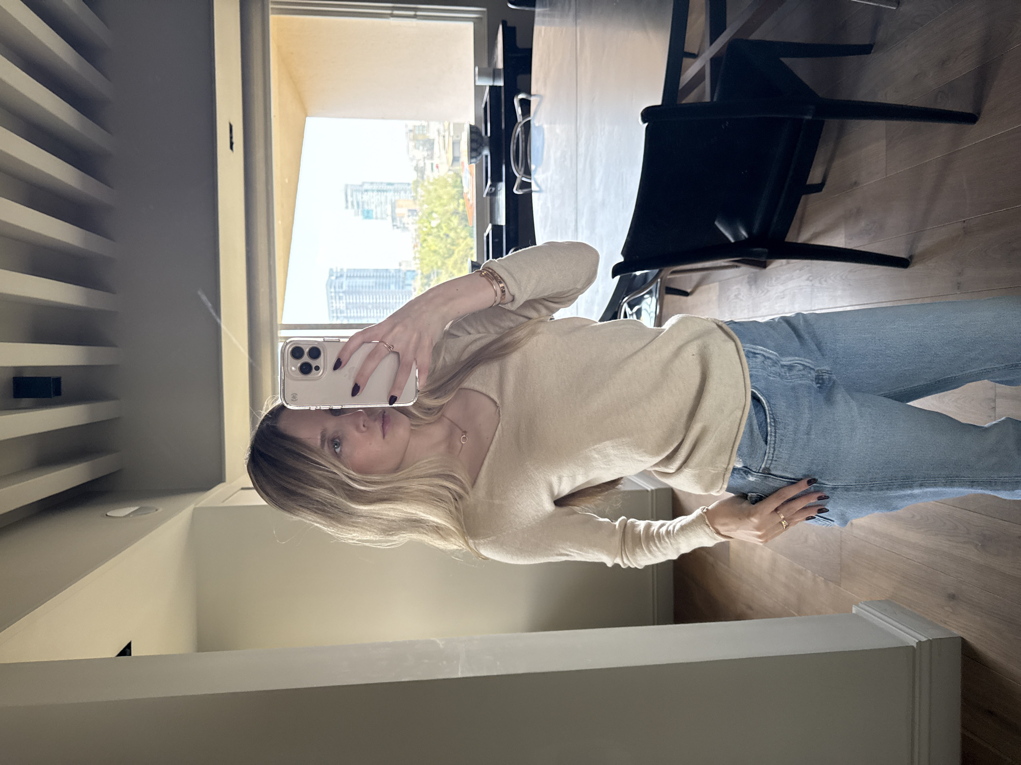 A young woman is taking a selfie in a mirror. She is wearing a light beige long-sleeved shirt and light blue jeans. She is holding a phone in her left hand. The mirror is in a room with hardwood floors and a white baseboard. In the background, there is a window with a view of a city skyline. It's unclear if this location is in Mexico City, Mexico. A black chair sits behind her and the table behind the chair suggests a dining room setting. Shelving is visible on the left wall of the mirror.