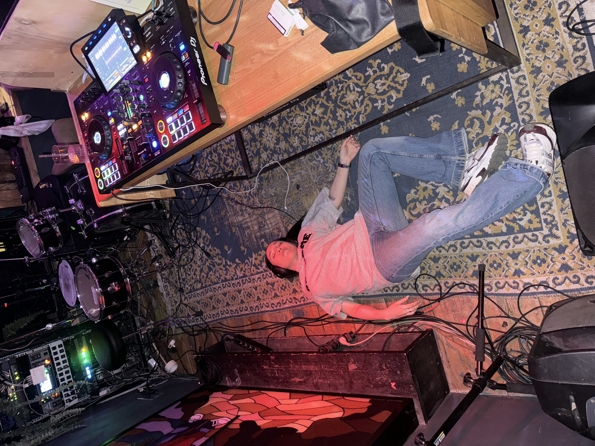 An individual lies supine on a patterned rug within an indoor music venue in Moscow, Russia. The person, wearing a light grey "SUPREME" t-shirt, blue jeans, and white/maroon athletic shoes, has headphones covering their ears. Their eyes are closed, and mouth is slightly open. The scene is illuminated by dim ambient light, red light from a wall art piece, and various colored LEDs from electronic equipment. To the left, a multi-tiered rack holds audio processing equipment with illuminated green indicators, positioned next to a black drum kit and stool. A wooden table supports a Pioneer DJ controller, actively displaying waveforms on its screen, alongside a glass containing a drink and straw, a black pouch, and a white cigarette pack. The floor is covered by a blue and yellow/beige patterned rug, extensively entangled with numerous black and white audio and power cables. A black speaker and microphone stands are visible in the foreground.