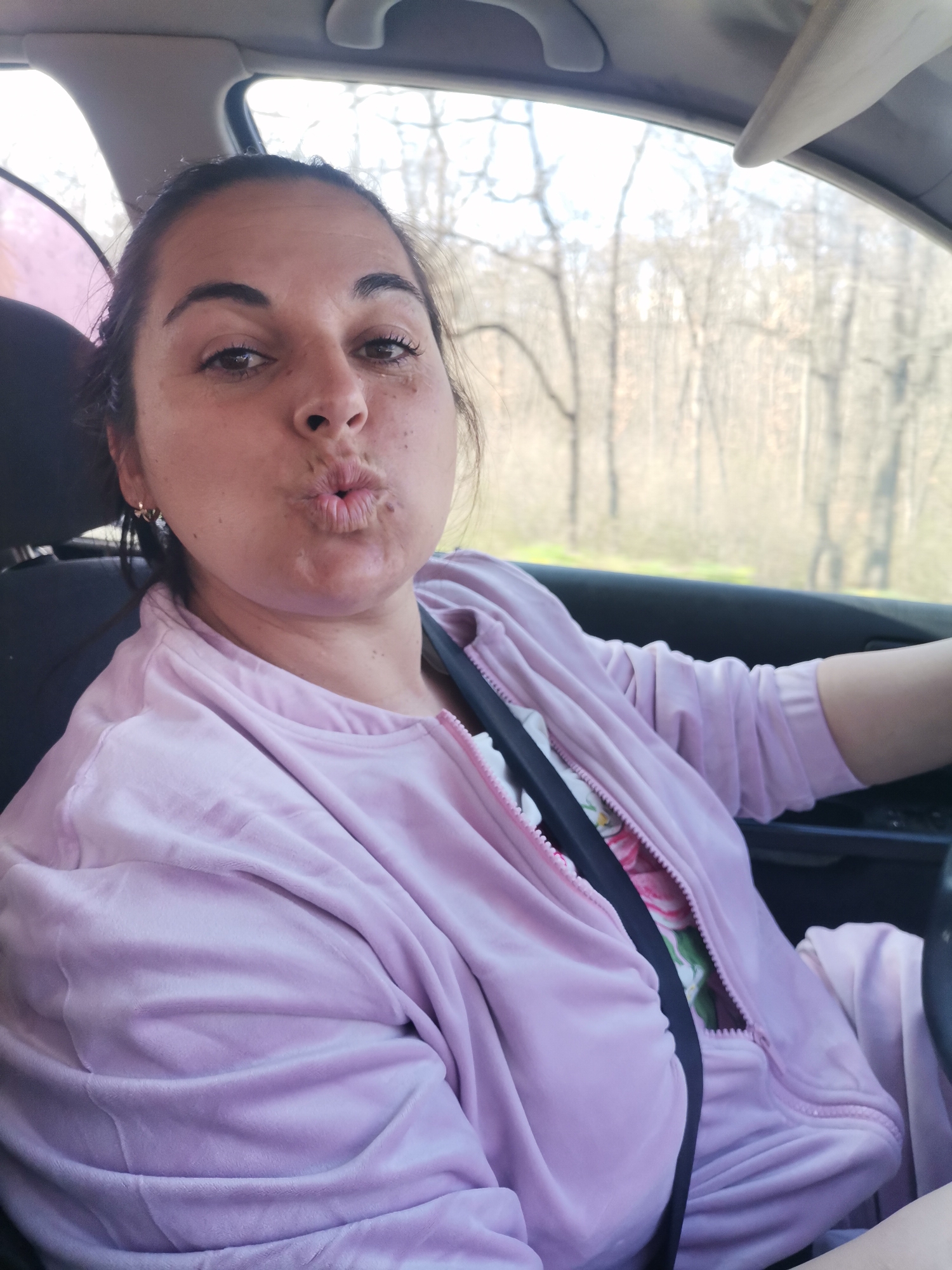 A woman is in the driver's seat of a car. She is wearing a pink velour tracksuit over a floral shirt and a seatbelt, and has her lips pursed as if to give a kiss. Outside the car windows is a view of bare trees. The picture was taken near Tsarkva, Bulgaria.