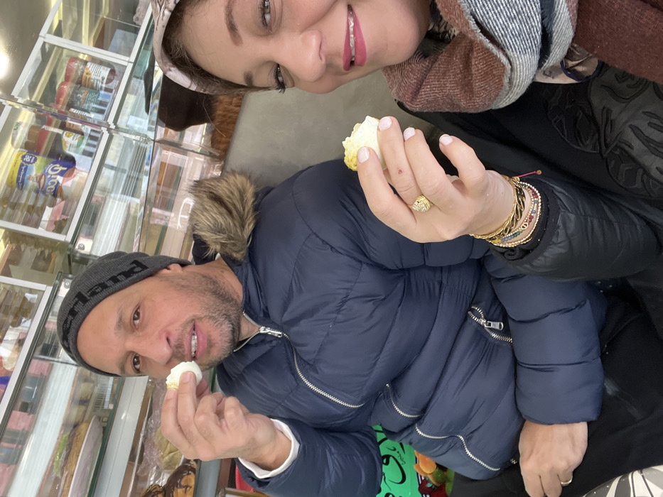 Two individuals, a male and a female, are depicted in a mid-shot within what appears to be a bakery or small grocery store in Meknes, Morocco. The male, positioned on the left, is approximately 40-50 years old, wearing a grey beanie with "PALACE" text, and a dark blue padded winter jacket featuring a fur-lined hood. He is holding a piece of a boiled egg in his right hand, elevated towards his mouth, and looks towards the camera with a slight smile. The female, positioned on the right and closer to the foreground, is also approximately 40-50 years old, wearing a light-colored headscarf, a dark garment, and a multi-toned scarf (brown, grey, white). She holds a piece of a boiled egg in her left hand, which exhibits a gold ring and multiple bracelets, and smiles broadly directly at the camera. The background features glass display cases containing various products: packaged dairy items, jars, and other goods on the upper shelves, and baked goods, including pastries and eclairs, on the lower shelves. Partially visible green signage with white Arabic calligraphy is present in the lower-left background. The scene is brightly illuminated.