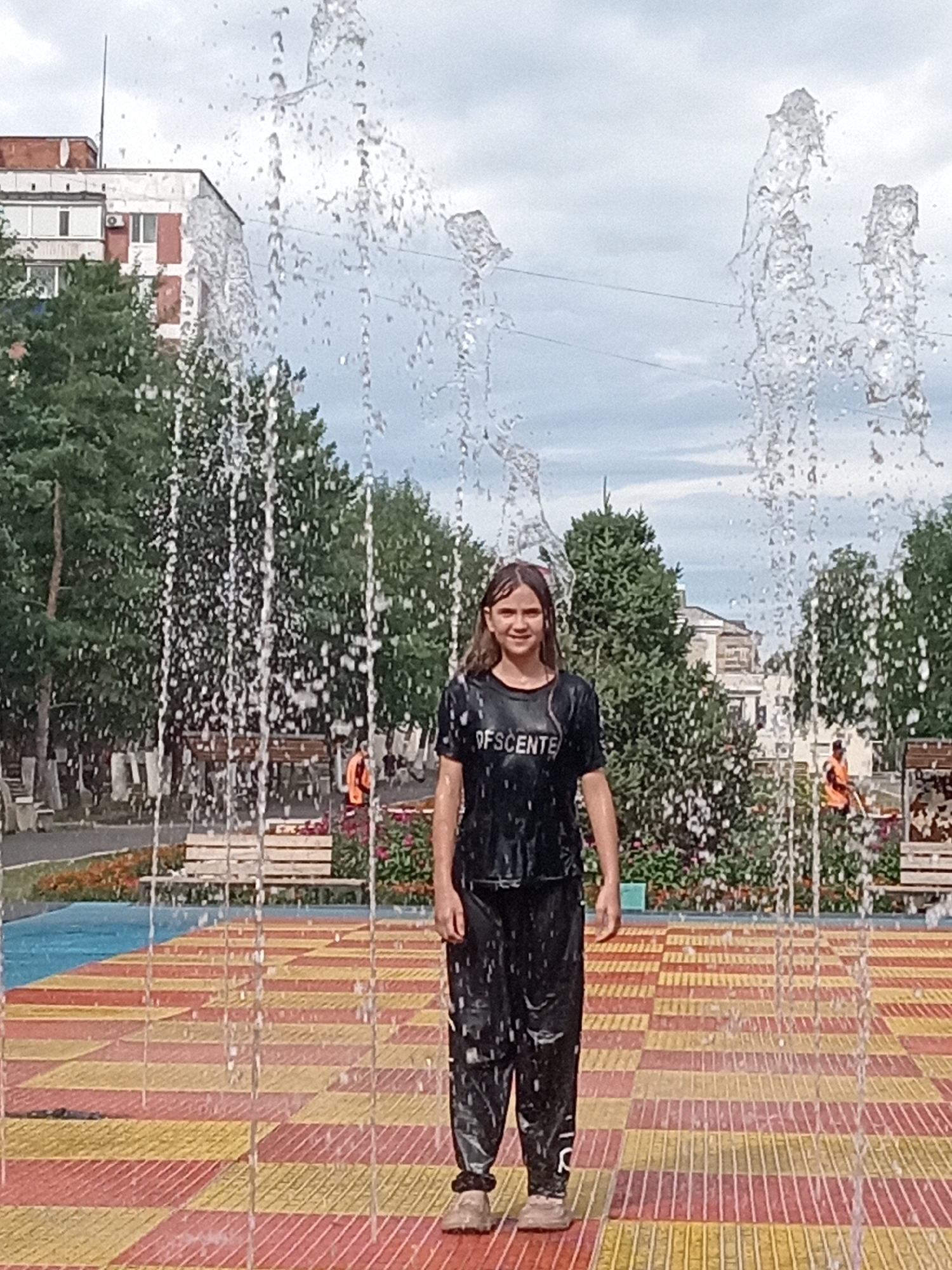 A young girl with brown hair, wearing a wet black t-shirt and black pants, stands in a splash pad. The t-shirt has "OFSCENTE" printed on it. She is smiling and looking at the camera. She is surrounded by water jets from a fountain. The splash pad has a red and yellow tiled surface. In the background, there are trees, buildings, and benches. The sky is cloudy. This scene is located in Rudny, Kazakhstan.