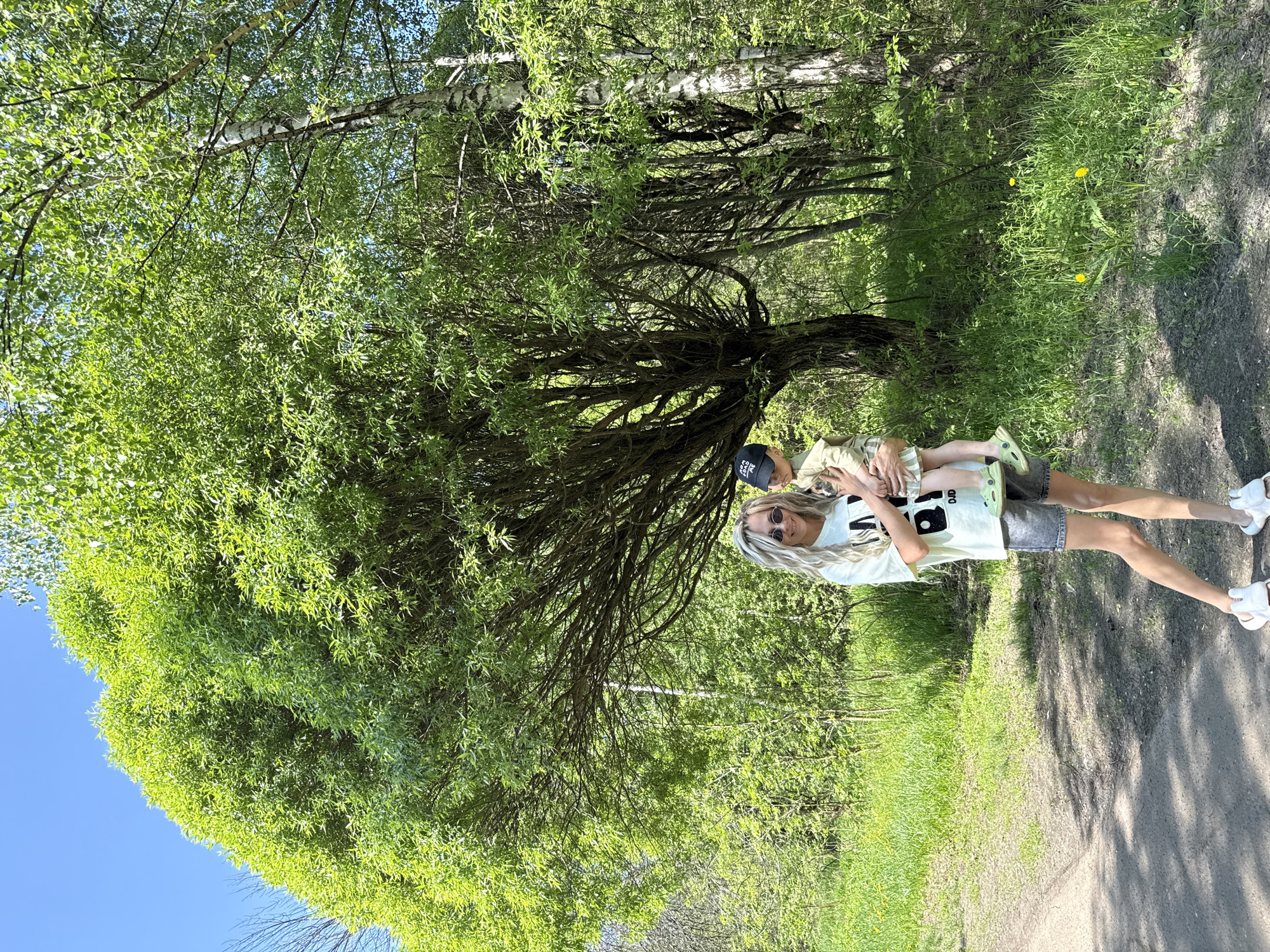 A woman is holding a child in a park-like setting, possibly Rusanovo, Russia, based on the location. The woman is wearing sunglasses and a light-colored t-shirt, and denim shorts. The child is wearing a baseball cap. They are standing on a path. The background includes trees and green foliage. The lighting suggests it is daytime.
