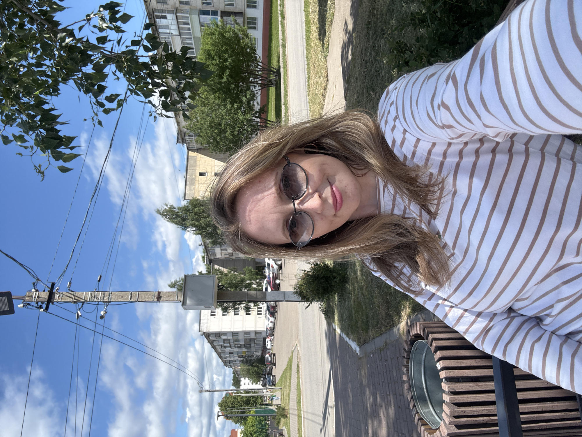 A woman with blonde hair and sunglasses is taking a selfie outdoors. She is wearing a long-sleeved white shirt with brown stripes. She is sitting on a bench, and a trash can is to her left. In the background, there are apartment buildings, trees, a street with cars, and a utility pole with wires. The sky is blue with some clouds. This appears to be a residential area in Chaikovsky, Russia.