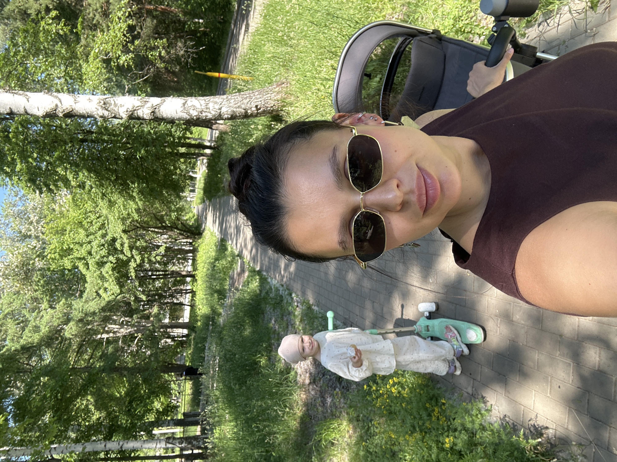 A woman, likely in her late 20s to early 30s, with dark hair pulled into a bun and wearing sunglasses and a dark brown sleeveless top, takes a selfie in a park. To her left, a young child, appearing to be around 3-4 years old, dressed in a cream-colored outfit and cap, stands on a teal scooter and holds a pastry. Behind the woman and to her right is a dark gray stroller. The scene is set on a paved path lined with green grass and trees, suggesting a sunny day in a park or a similar outdoor recreational area, possibly in the city of Chaikovsky, Russia, given the environment depicted.