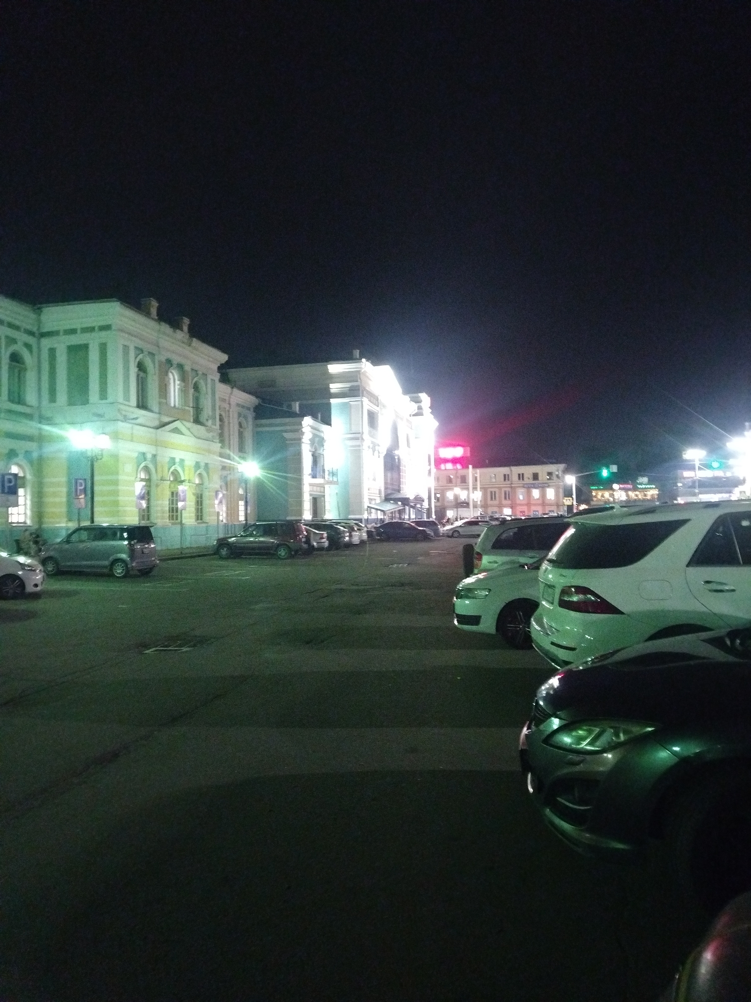 A nighttime outdoor street scene in Irkutsk, Russia, depicts a parking lot in front of a historical building with arched windows and ornate architecture, illuminated by streetlights. Several cars are parked, including a silver minivan, a dark-colored SUV, a white SUV, and a dark sedan in the foreground. The building appears to be a station or public facility, with a pedestrian crossing visible in the background. The sky is dark, indicating it is nighttime.